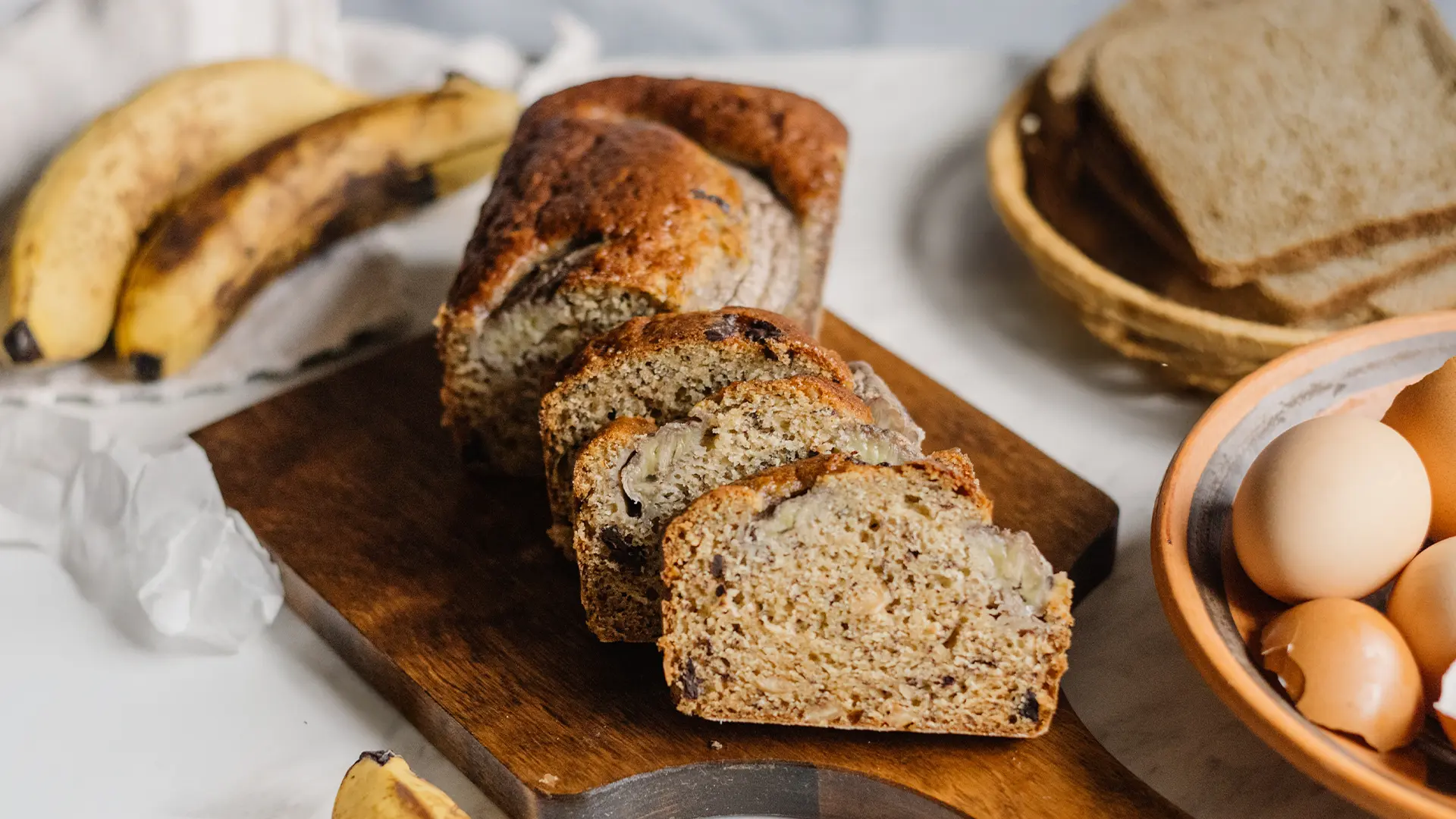 Freshly baked banana bread loaf sliced on a wooden cutting board, with bananas, eggs, and bread slices in the background