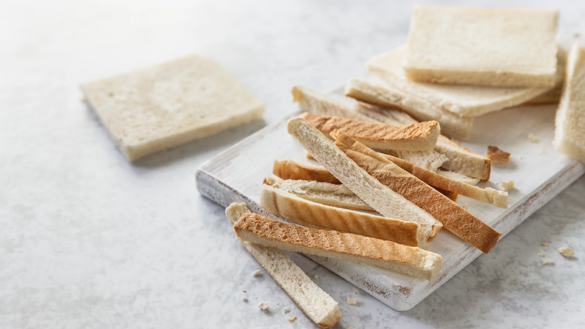 Slices of Hovis Farmhouse Soft White bread with the crusts removed, placed on a cutting board with the crusts set aside, in a marbled background. 