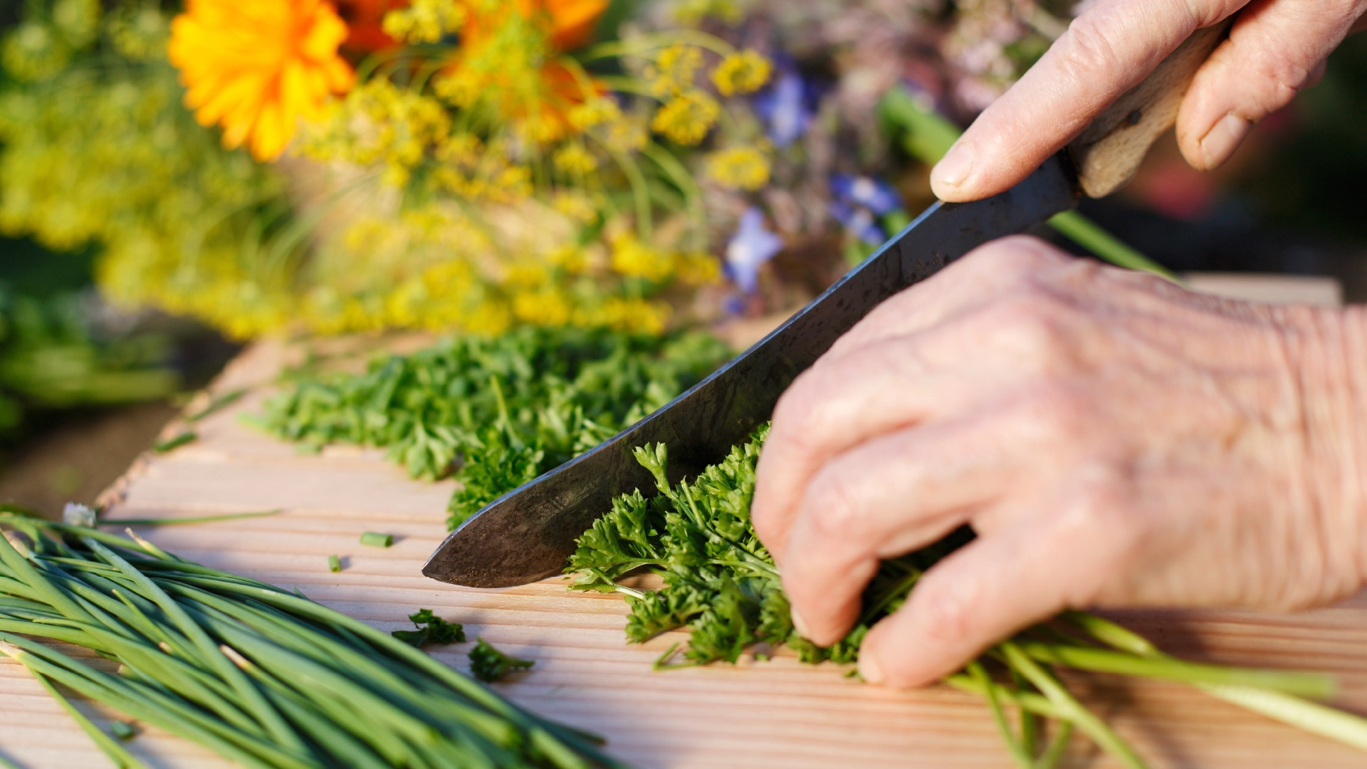 A hand holds down some herbs on a wooden chopping board while the other uses a sharp knife to chop them. In the background, some flowers and greens.