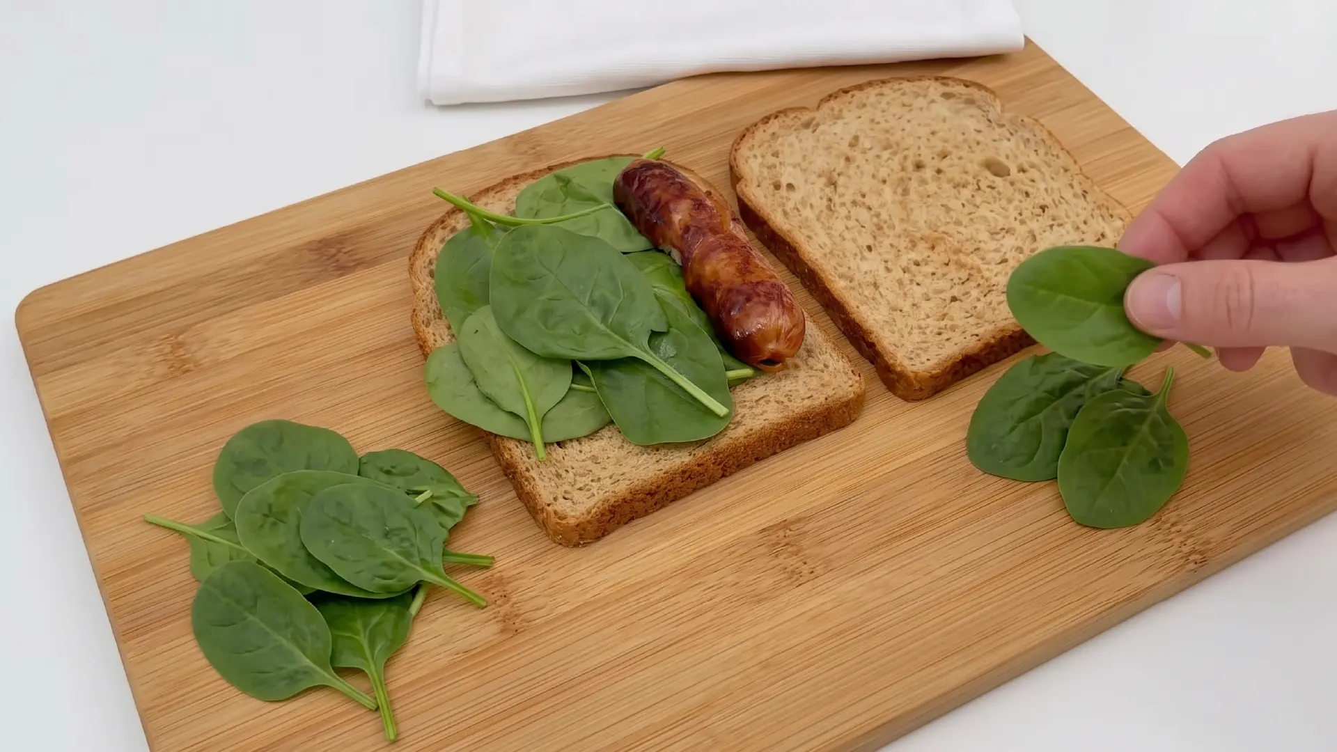 A hand adding fresh spinach leaves to a sausage sandwich on a wooden cutting board with whole wheat bread.