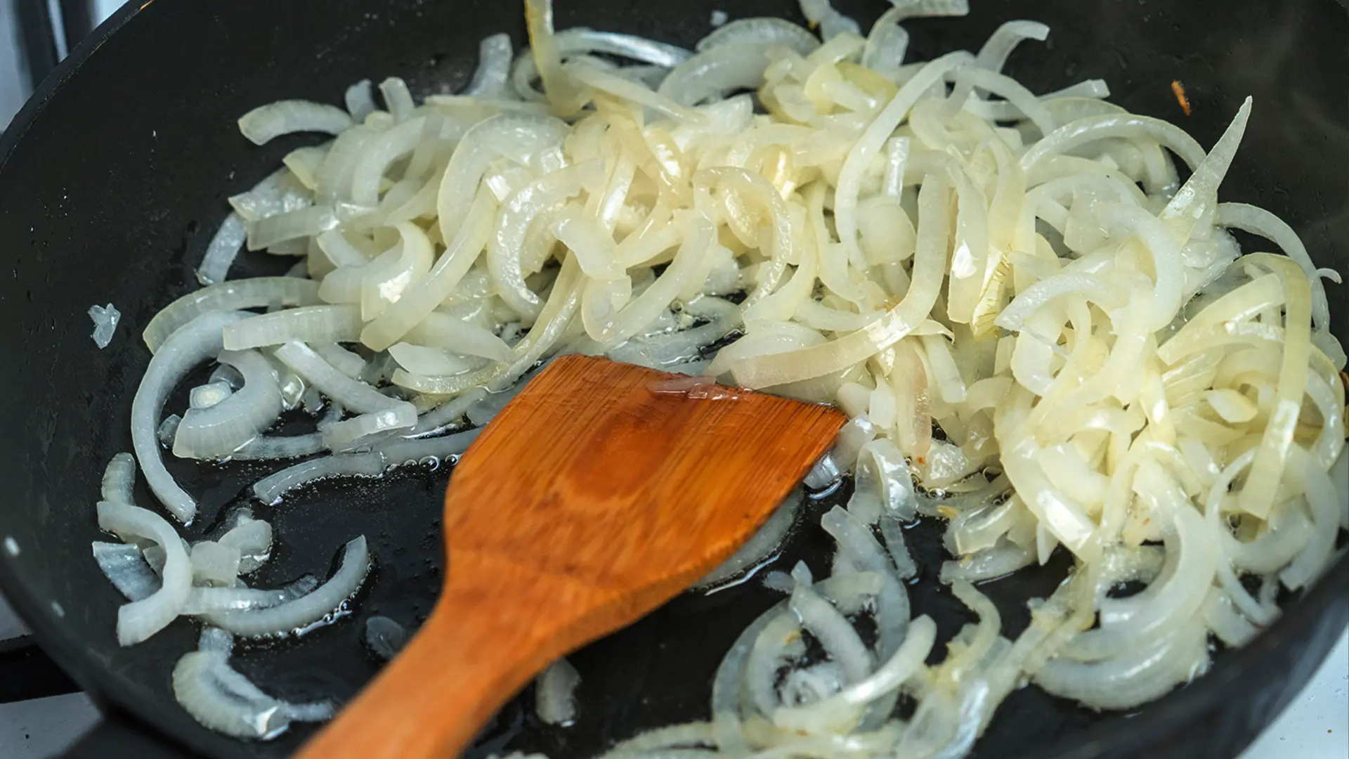 Thinly sliced white onions being sautéed in a dark frying pan. A wooden spatula is in the foreground, stirring the translucent onion slices. Some of the onions are beginning to caramelize slightly. The pan is on a stove burner, and the overall scene suggests the initial step of cooking.