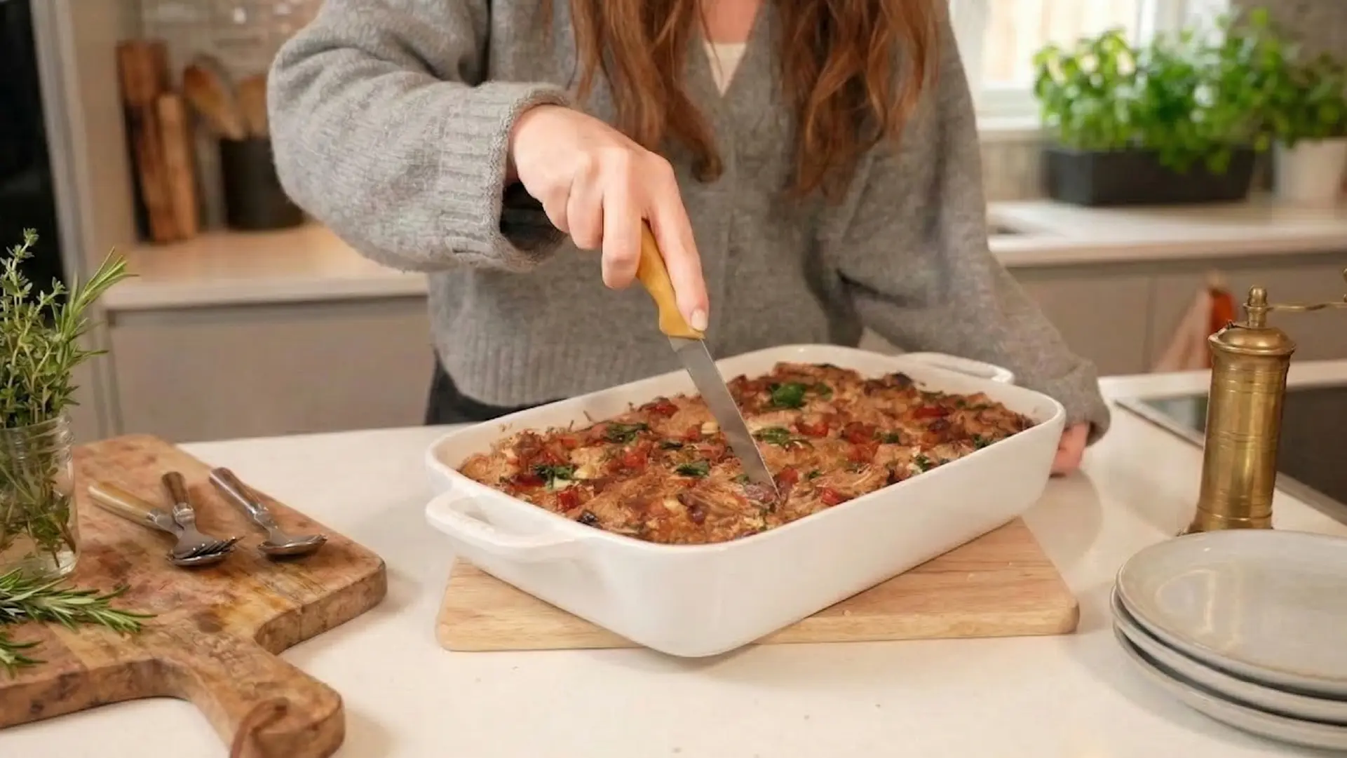 A person slices a golden-brown, baked breakfast strata in a white dish using a wooden-handled knife in a bright kitchen.