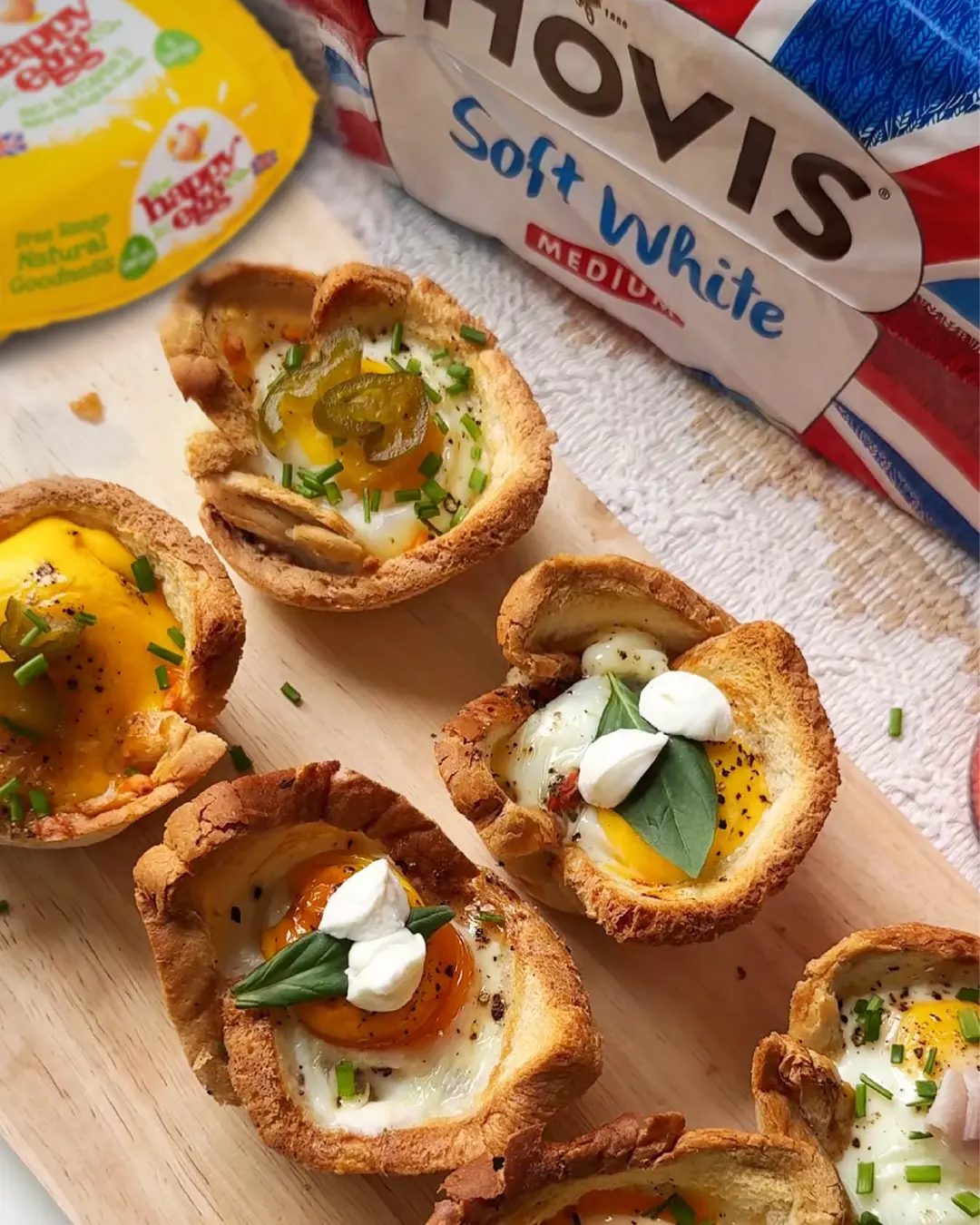 Baked toast cups filled with eggs, herbs, and toppings next to Hovis Soft White bread and a Happy Egg Co. carton.