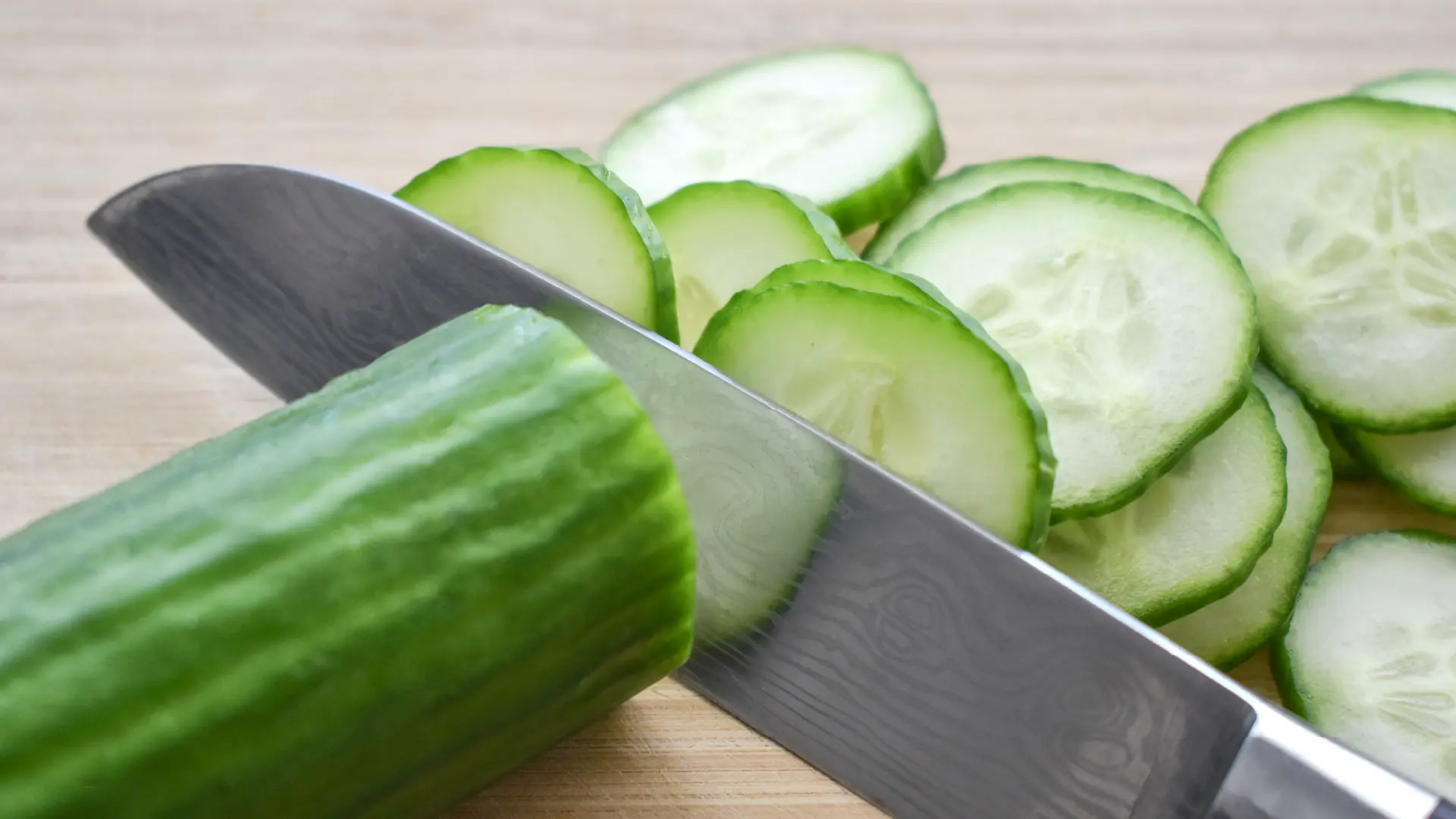 A sharp steel chef's knife slicing a fresh green cucumber into round pieces next to a pile of slices on a wooden board.