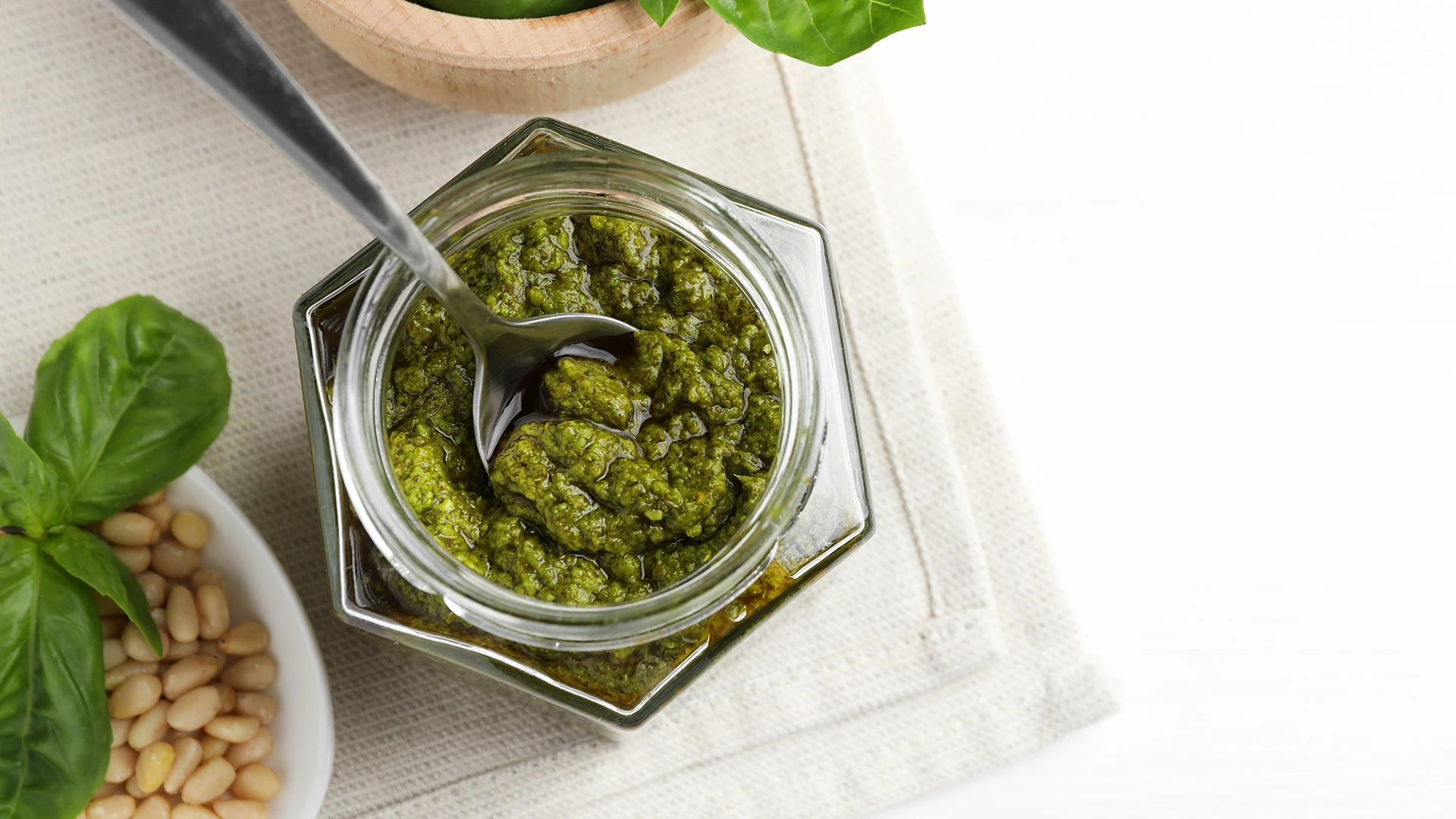 A top-down view of a hexagonal glass jar filled with green pesto and a spoon inside, with a small bowl of pine nuts and basil leaves on the side.