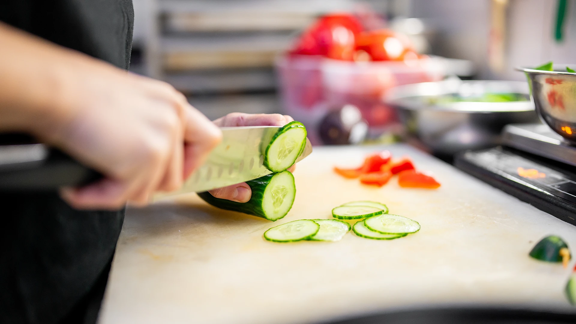 Hands using a sharp knife to thinly slice a cucumber on a wooden chopping board. In the background some tomatoes and greens in bowls.