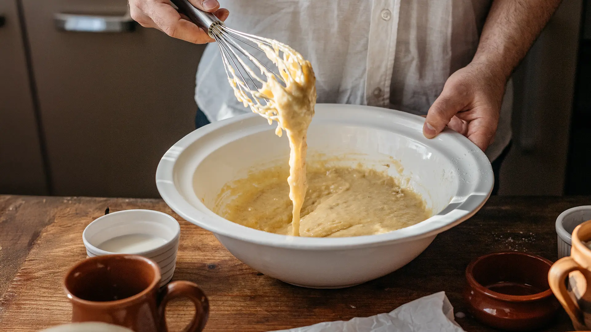 Person whisking banana bread batter in a large white mixing bowl, with the thick mixture dripping from the whisk