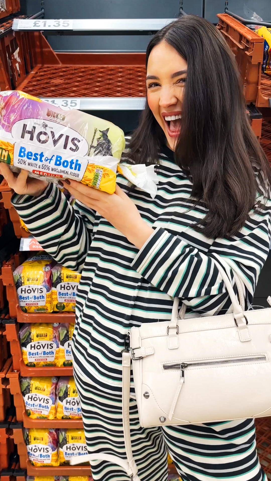 A cheerful woman stands in a grocery store aisle, smiling widely while holding a loaf of Hovis “Best of Both” bread. She is wearing a striped dress and carrying a cream-colored handbag on her arm. Her energetic pose and big smile give the scene a fun, enthusiastic feel, she’s excited about finding the product.