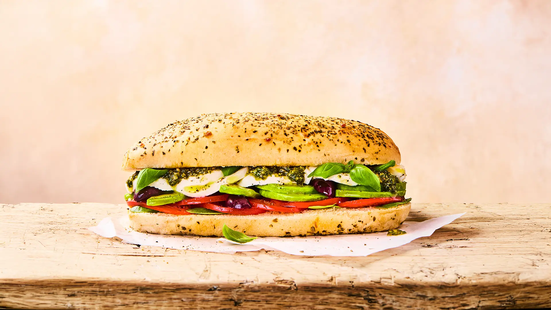 A fresh Hovis Cheese & Herb Sub Rolls sandwich, filled with pesto, sliced mozzarella, basil leaves, tomatoes, avocado and black olives rest on top of a napkin in a light beige background. 