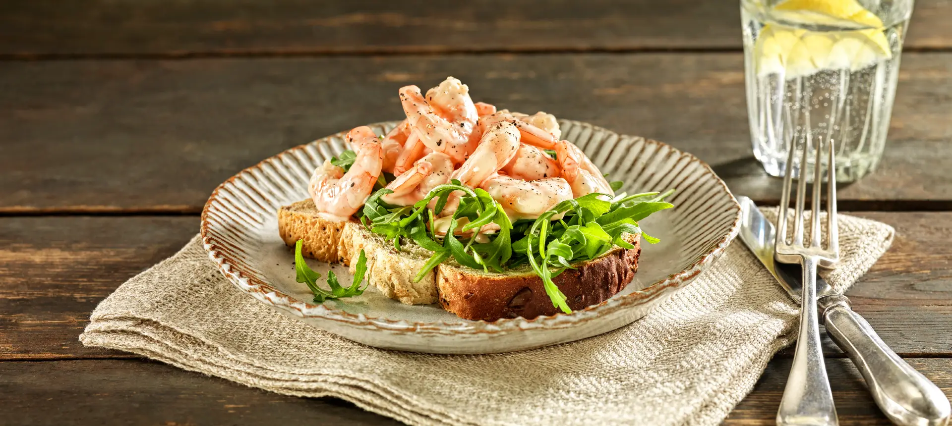 A slice of Hovis Seed Sensations Original 400g bread topped with rocket, prawns, and a sprinkle of ground black pepper, on a plate with cutlery on the side.