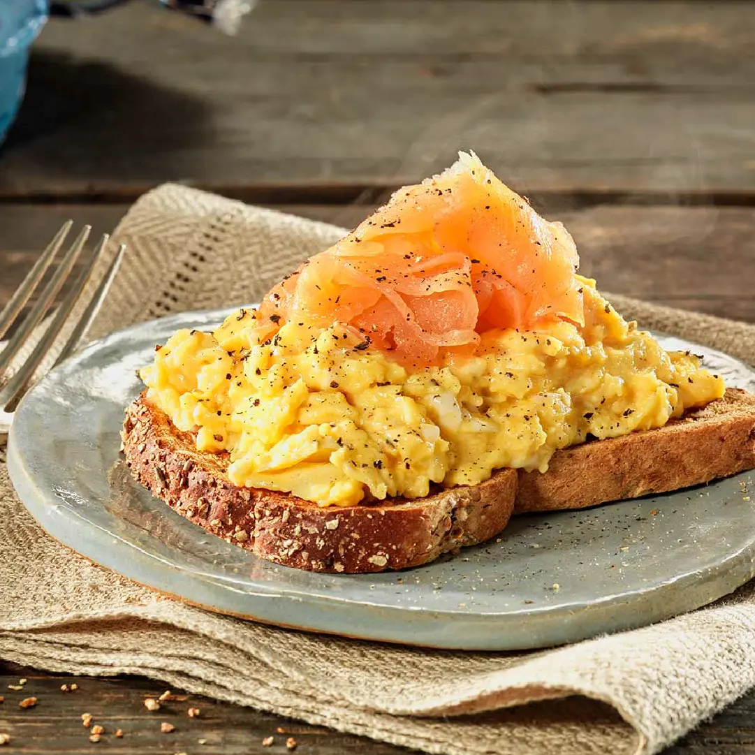 A slice of Hovis Granary Medium Sliced on a plate, topped with scrambled eggs, slices of smoked salmon, and a sprinkle of black pepper.