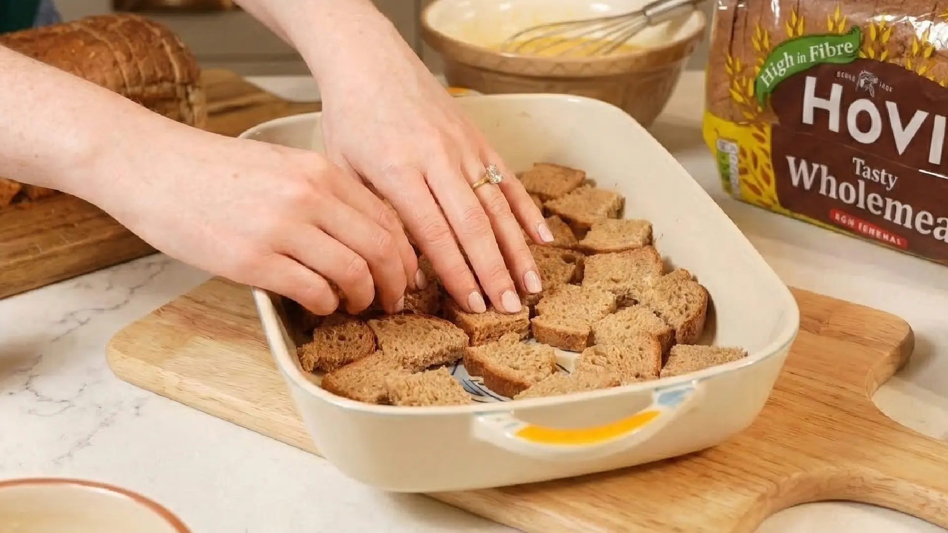 Hands layering cubes of wholemeal bread into a ceramic baking dish, with a Hovis bread loaf in the background.