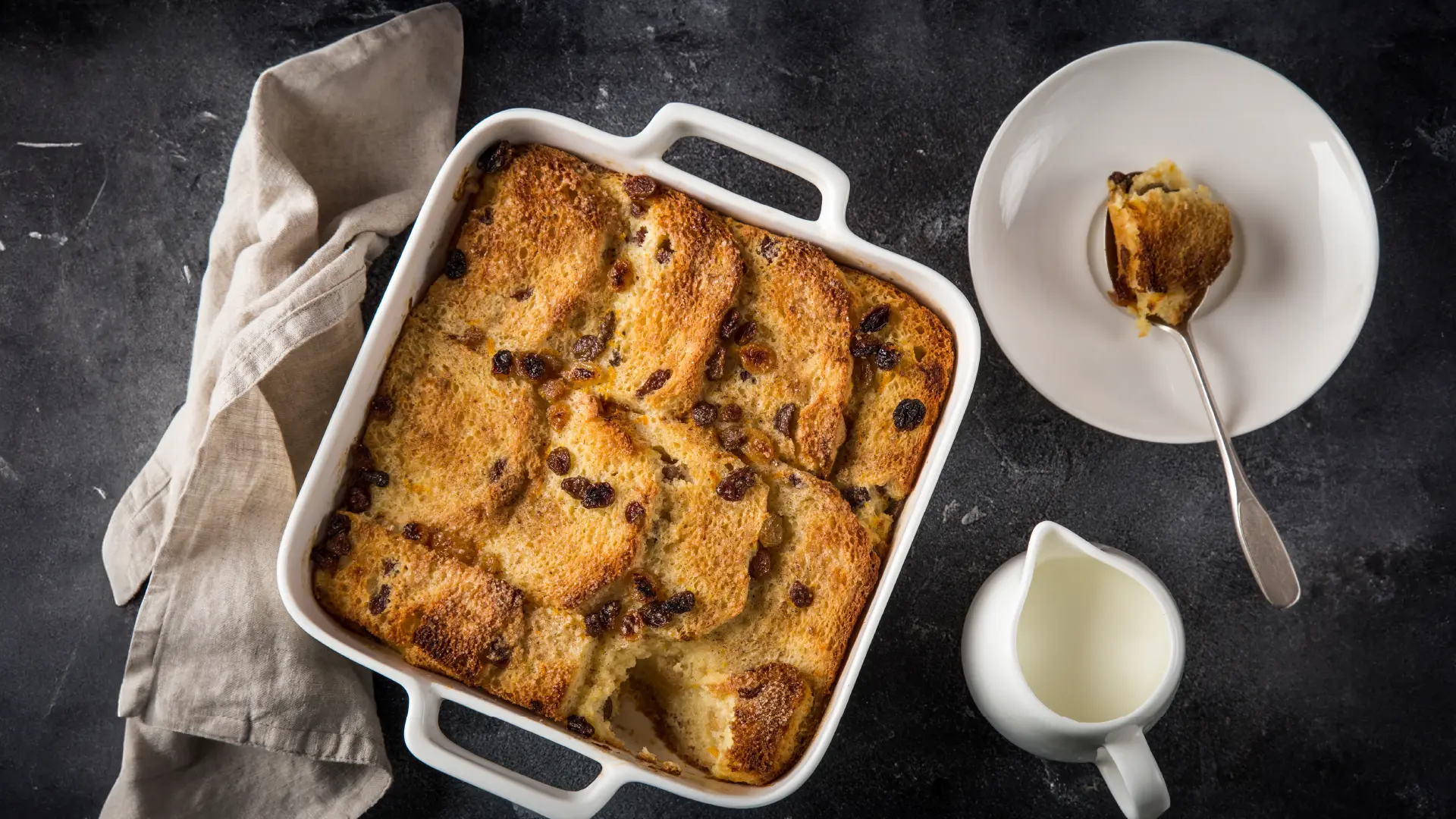 A top down view of a square baking tray with layered toasted Hovis Farmhouse Batch bread topped with raisins. Next to it, a small plate with a spoonful of bread pudding.