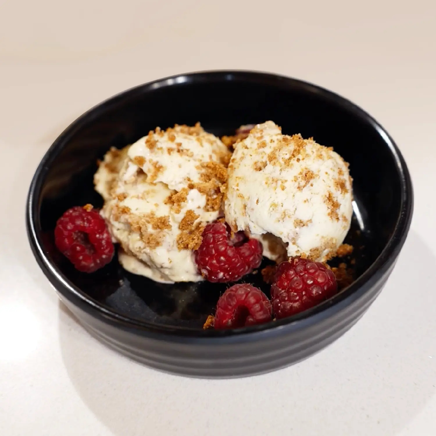 Two scoops of ice cream topped with crushed nuts and served with four fresh raspberries in a black bowl.
