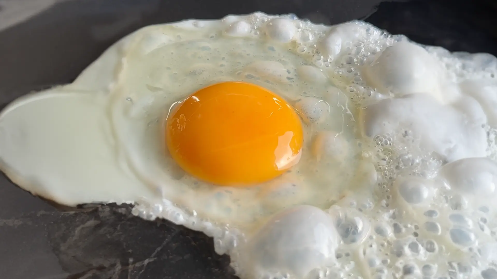 An egg being fried with oil in a black frying pan.