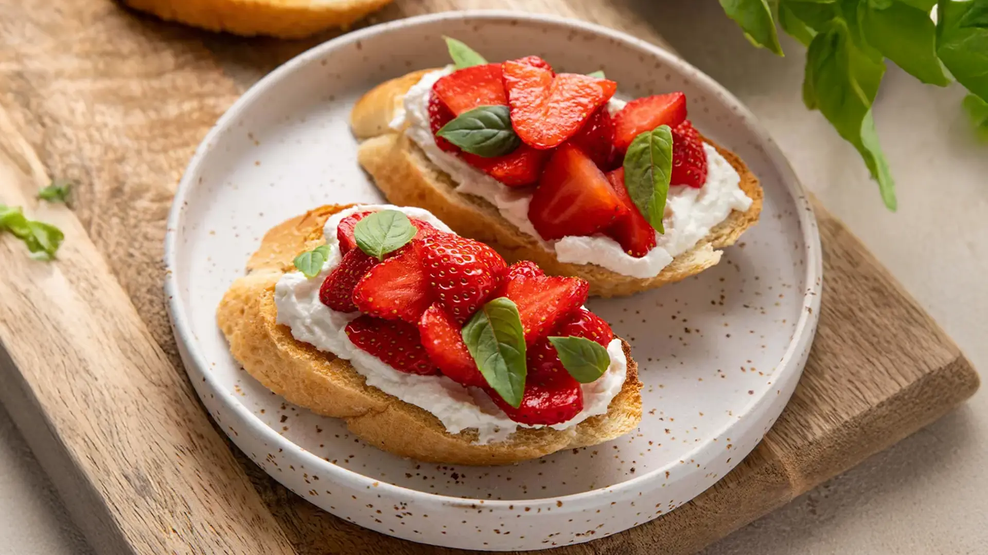 Two slices of toasted bread topped with creamy cheese, fresh strawberry slices, and basil leaves, served on a speckled ceramic plate.