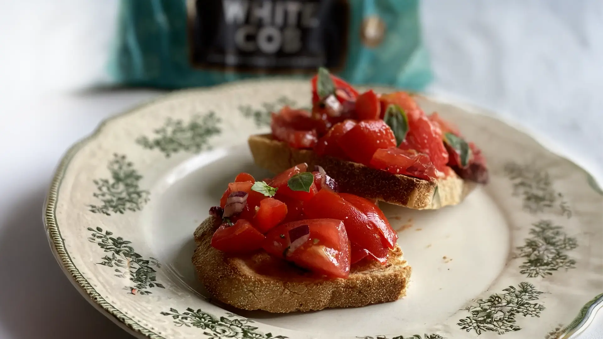 A close up of two half slices of Hovis White Cob on a floral plate, topped with a mix of diced tomatoes, onions and basil. In the background, a packaging of Hovis White Cob. 