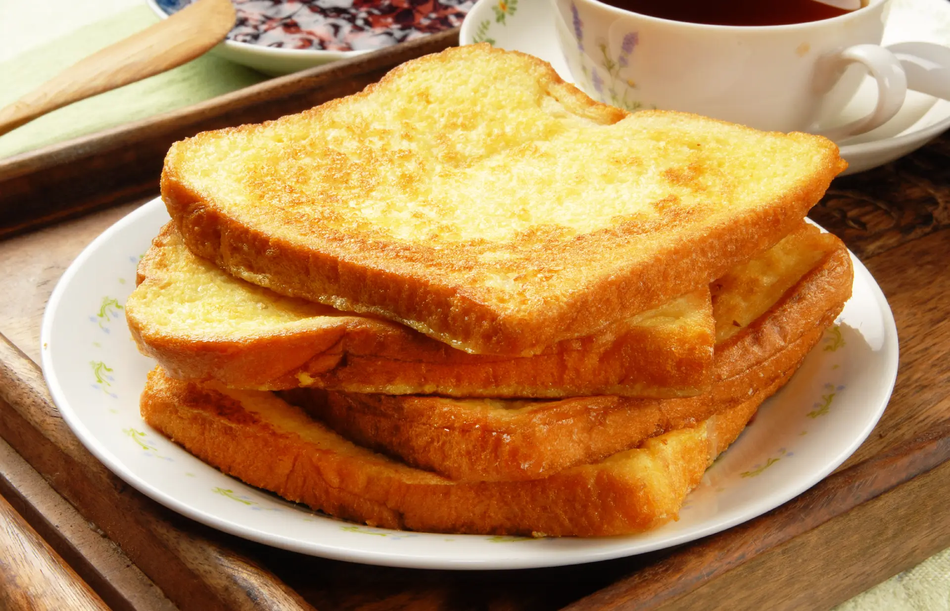 Four slices of toasted Hovis Soft White Extra Thick Sliced bread stacked on a plate, with a cup of tea and a small plate of jam on the side.