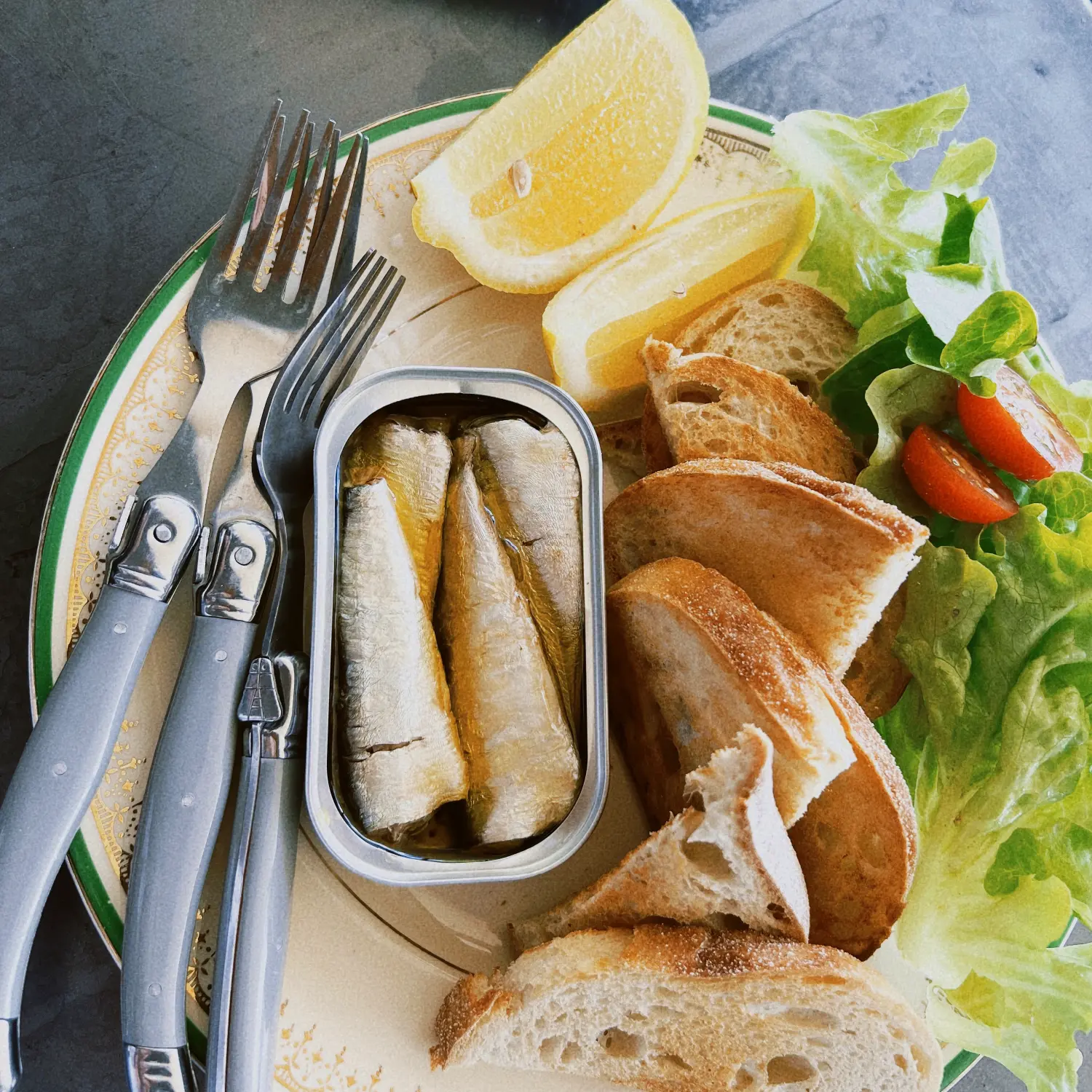 An open tin of sardines on a plate with sliced bread, lemon wedges, a fresh side salad and two silver forks.