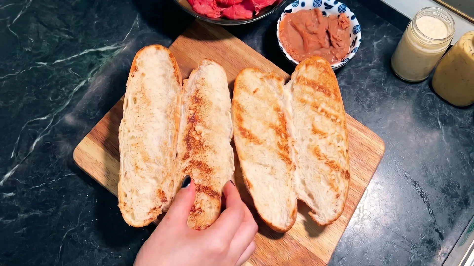 A hand arranges toasted, sliced baguettes on a wooden board, with bowls of spreads and jars nearby on a dark counter.