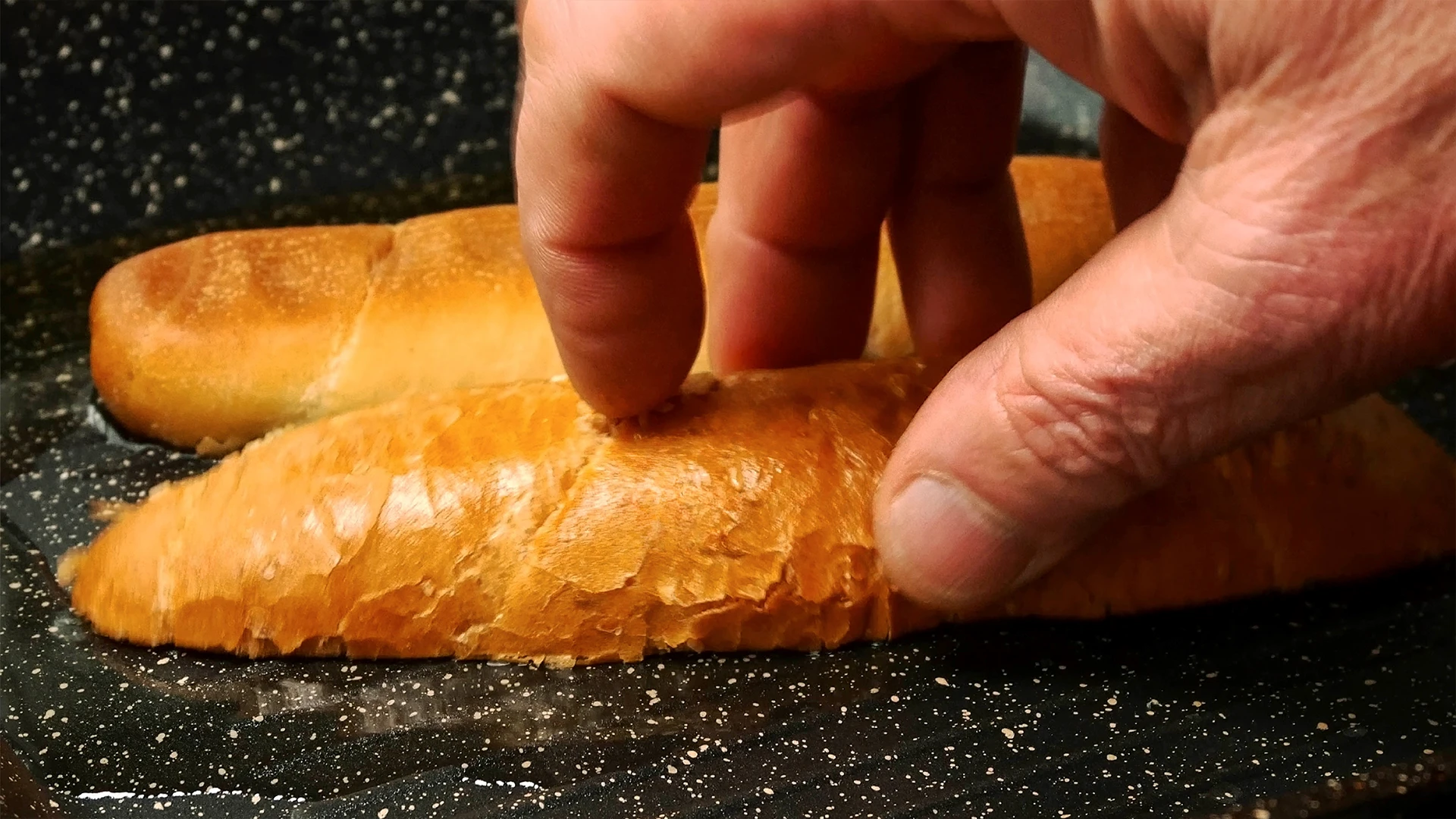 A hand holds a half sub roll on an oiled frying pan to toast