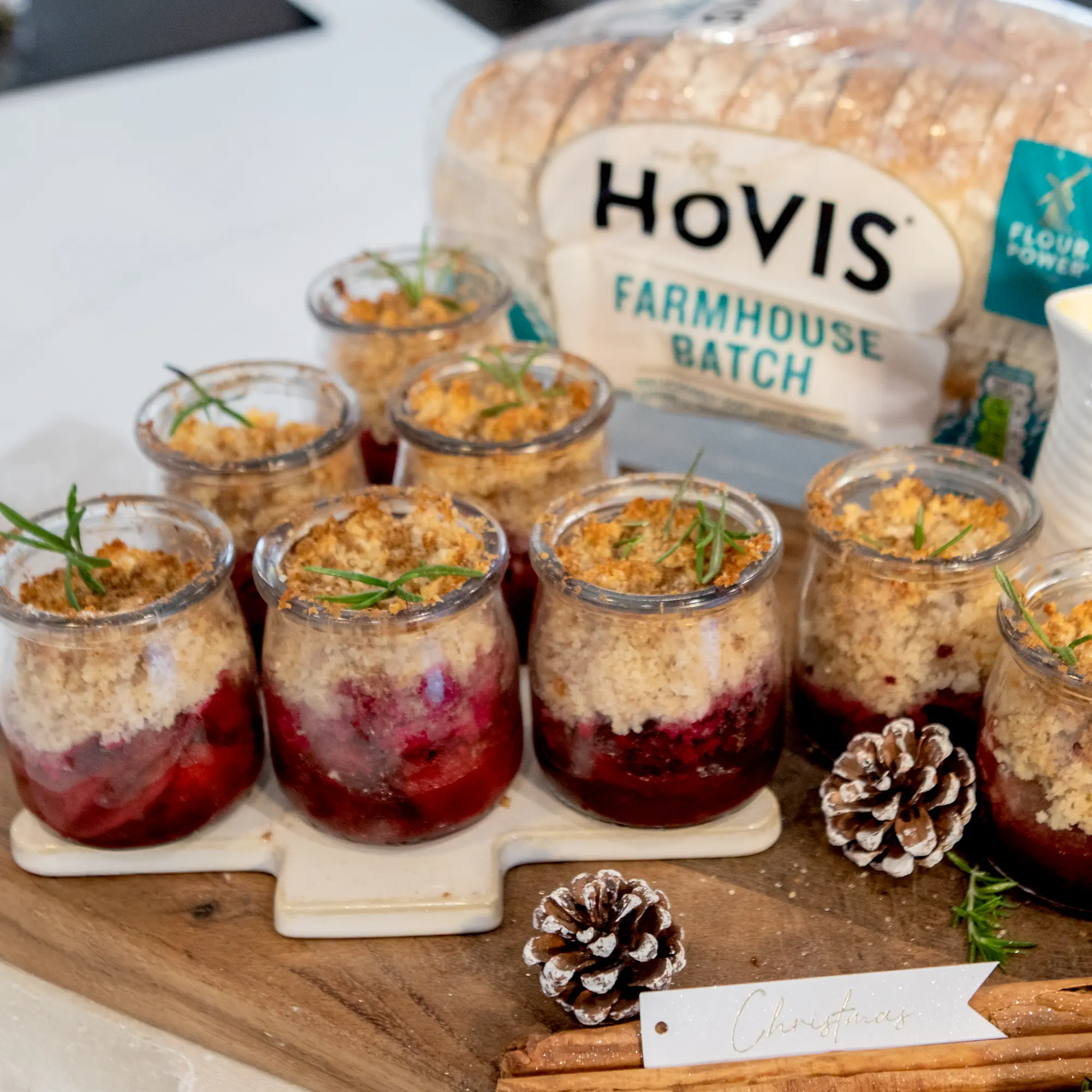 A close-up of eight small pots filled with golden fruit Christmas crumble, arranged on a wooden board decorated with pine cones and a festive “Christmas” badge. In the background, a loaf of Hovis® Farmhouse Batch sits on the counter, showing the bread used to make the crumble topping.