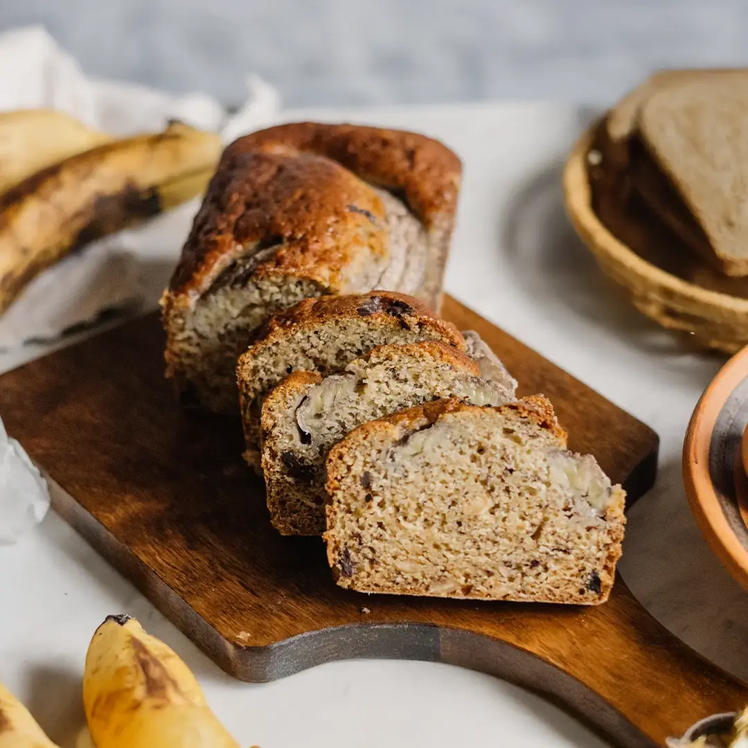 Freshly baked banana bread loaf sliced on a wooden cutting board, with bananas, eggs, and bread slices in the background