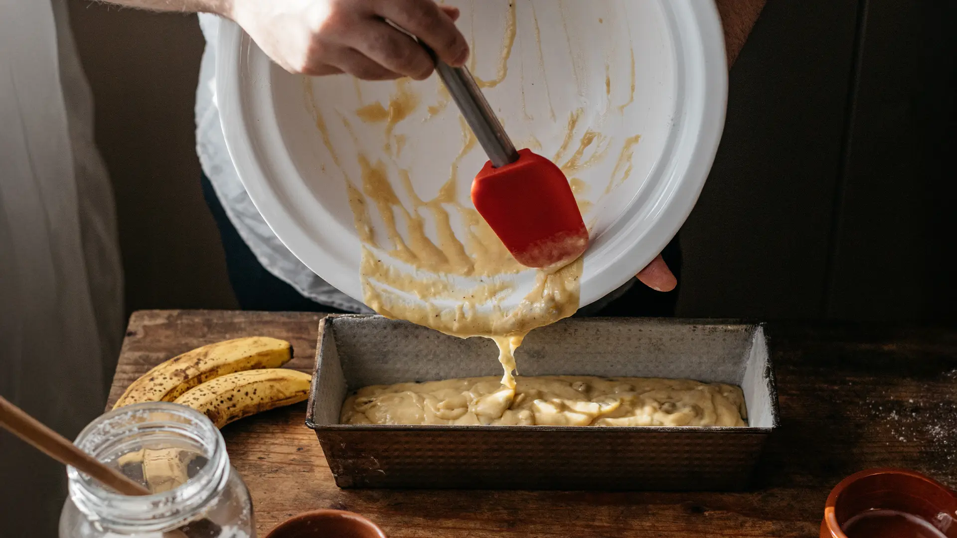Person pouring banana bread batter from a mixing bowl into a rectangular loaf pan using a red spatula