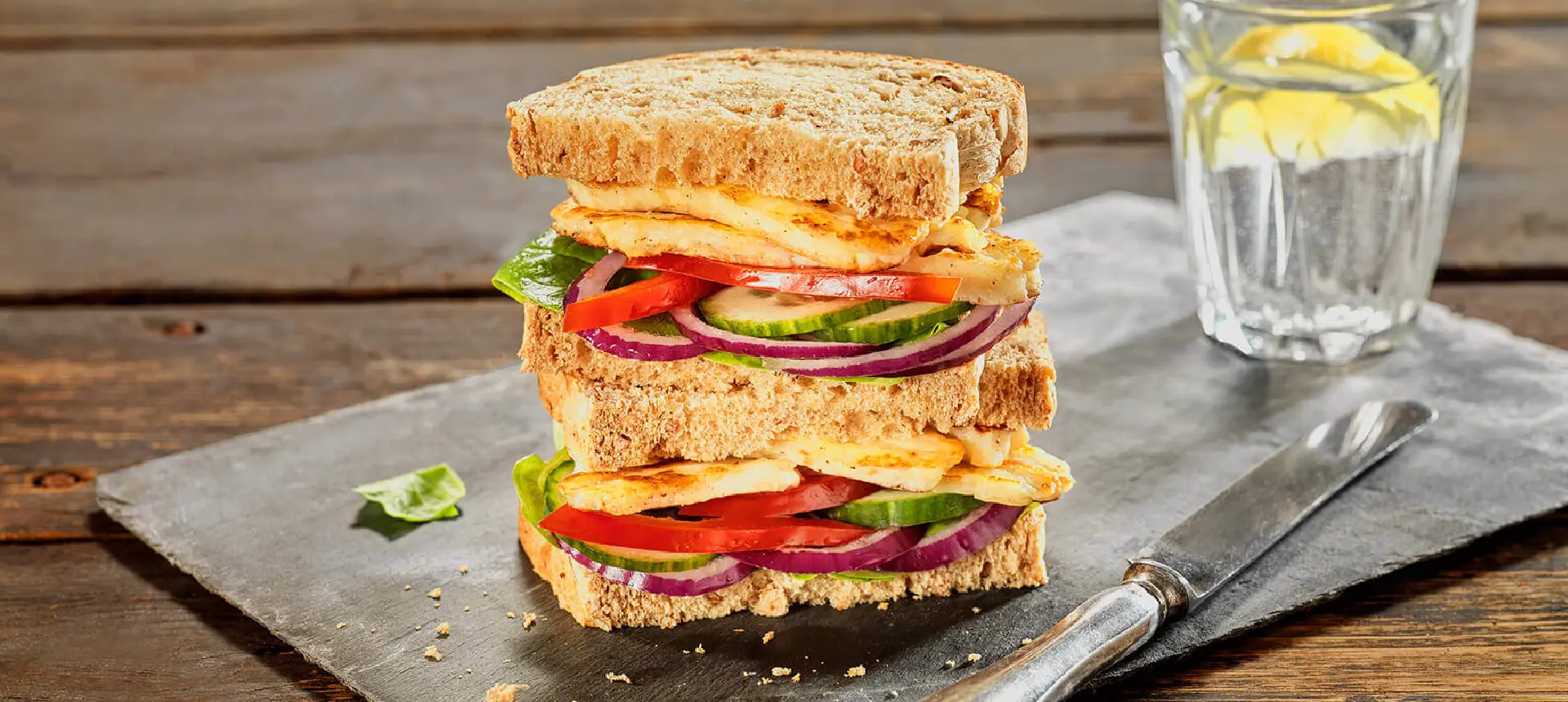 Two sandwiches made with Hovis Granary 400g stacked, filled with slices of red onion, cucumber, red pepper, and halloumi between brown bread slices.