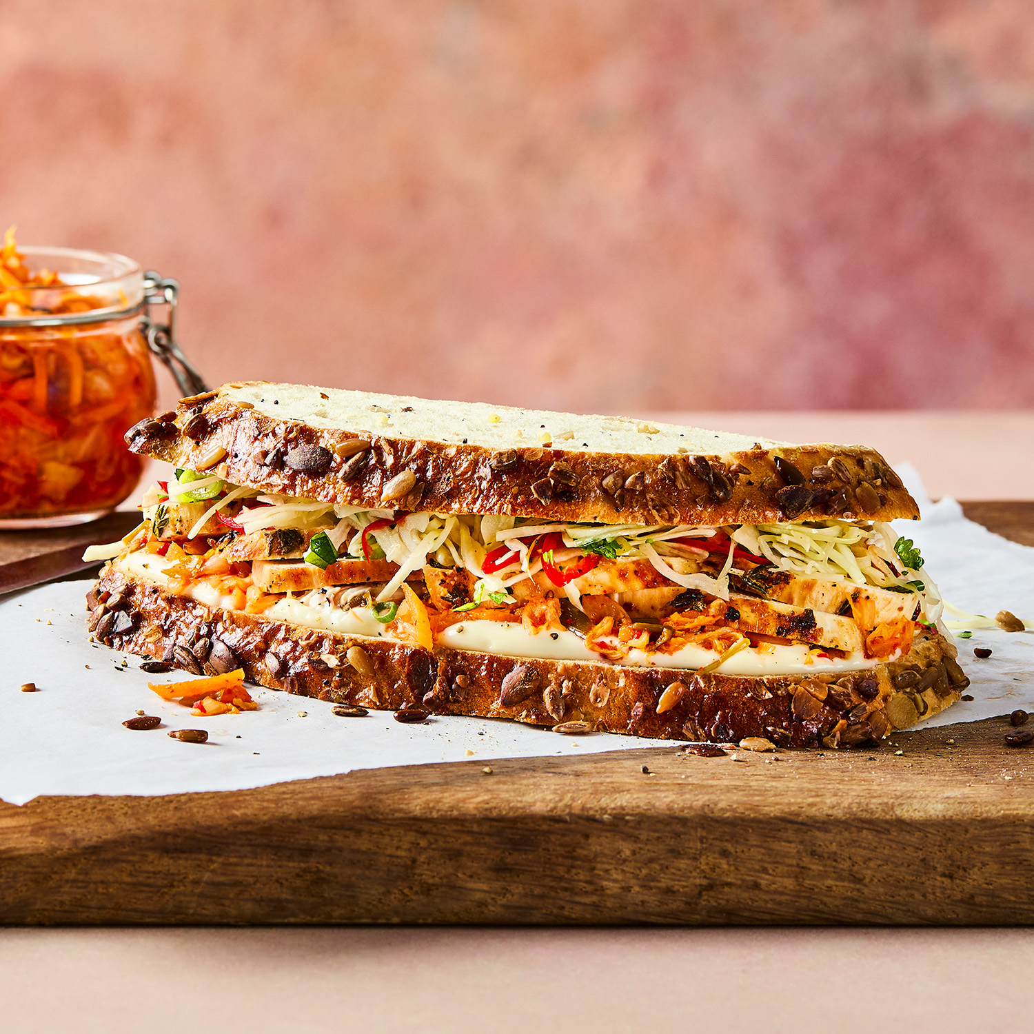 a seeded sourdough sandwich with slaw, kimchi and chicken breast pieces inside, sitting on a wooden chopping board with a bowl of leftover kimchi in the background 