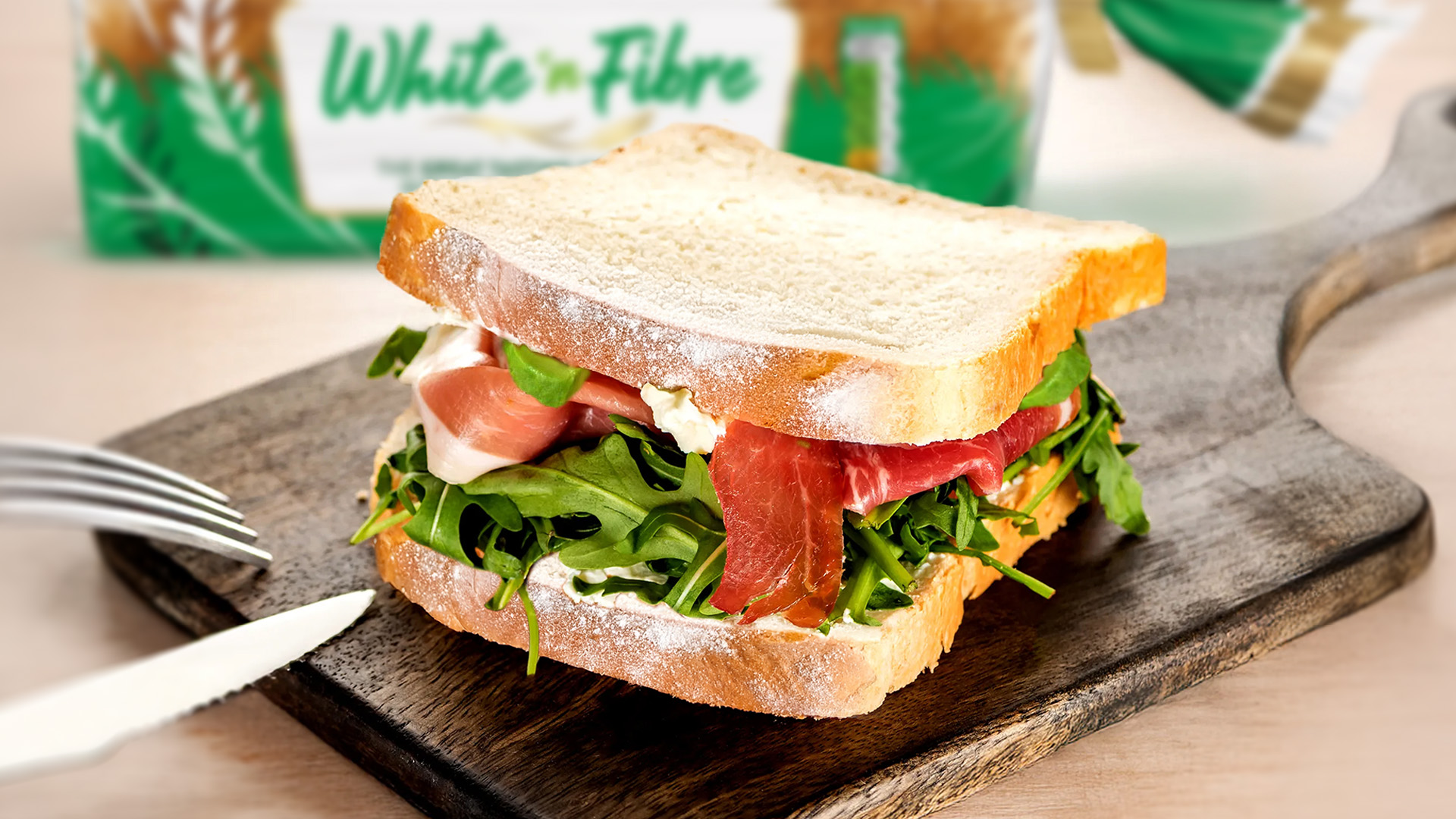 A sandwich made with Hovis White 'n Fibre bread sits on a dark wooden cutting board. The sandwich is filled with cheese spread, fresh rocket, and delicate slices of cured ham. The top slice of bread is slightly askew, revealing the delicious layers inside. A small portion of a knife and fork are visible to the left. In the background, a blurred loaf of Hovis White 'n Fibre bread in its green packaging adds context.
