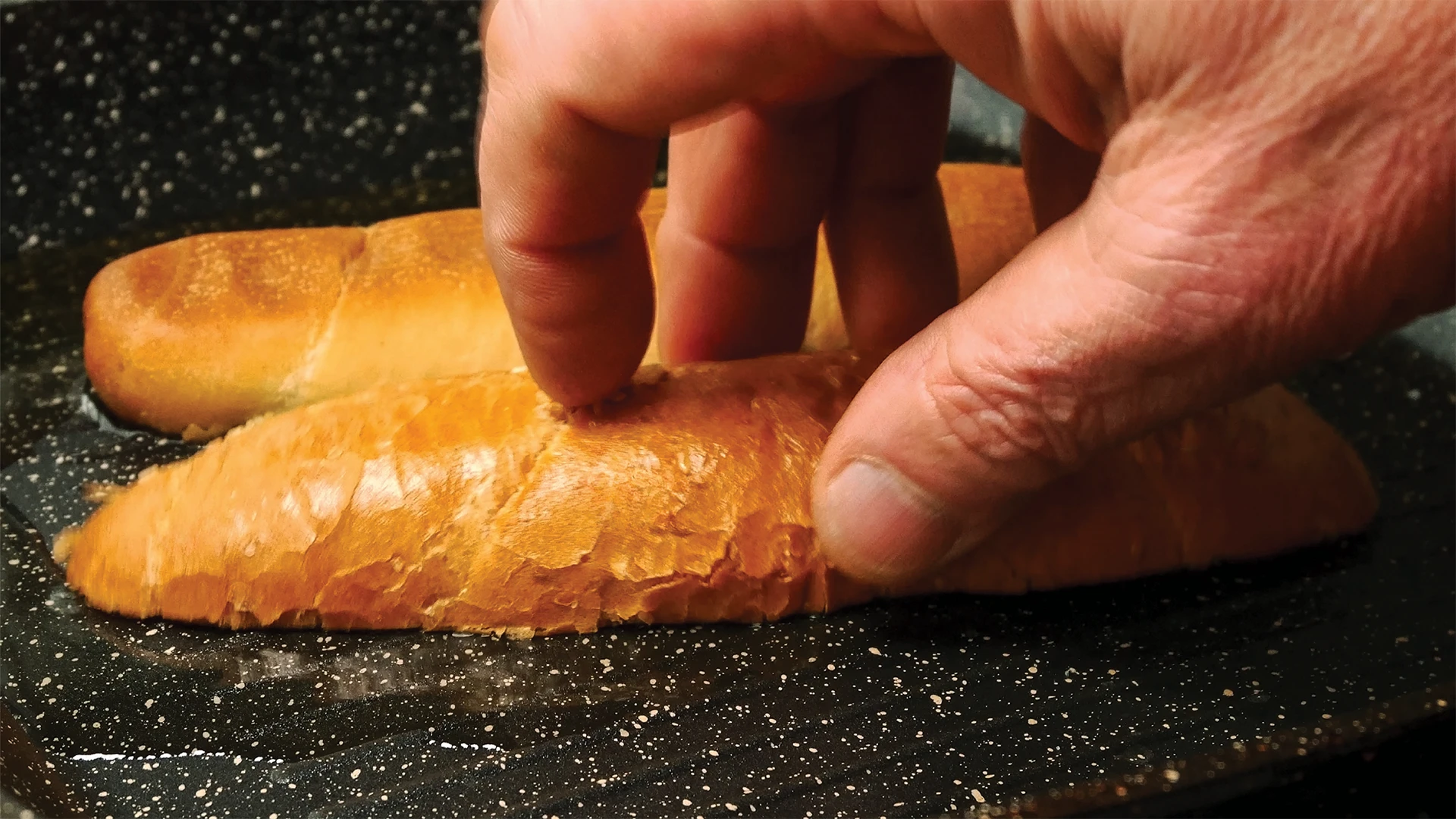 A hand holds a sub roll on a oiled frying pan to toast