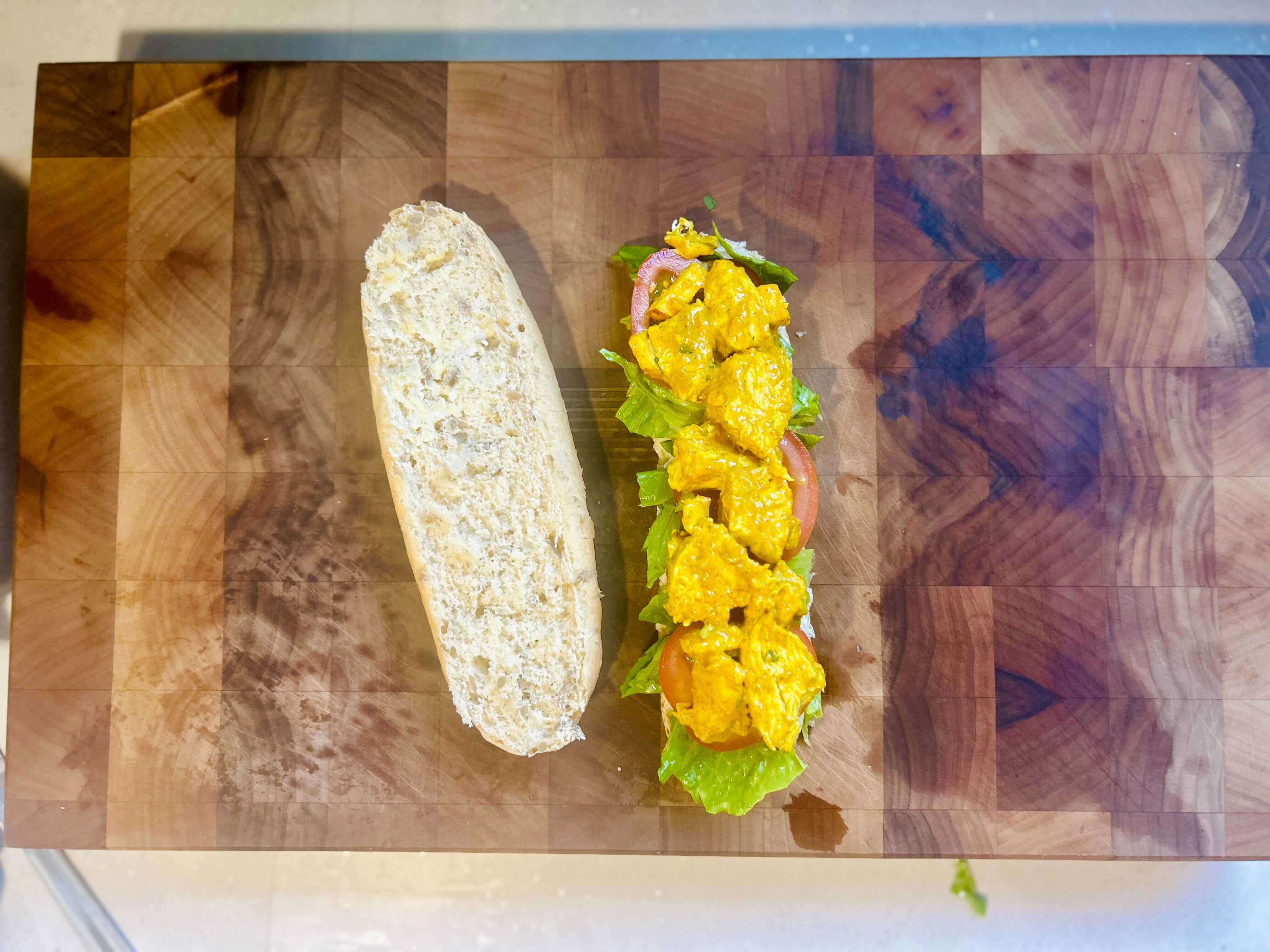 A top-down shot of a large, opened sub roll on a wooden chopping board. On the right side of the board is the bottom half of the roll, which is filled with layers of shredded lettuce, sliced tomatoes, and a portion of creamy yellow Coronation Chicken. The top half of the roll is sitting empty on the left side of the board.