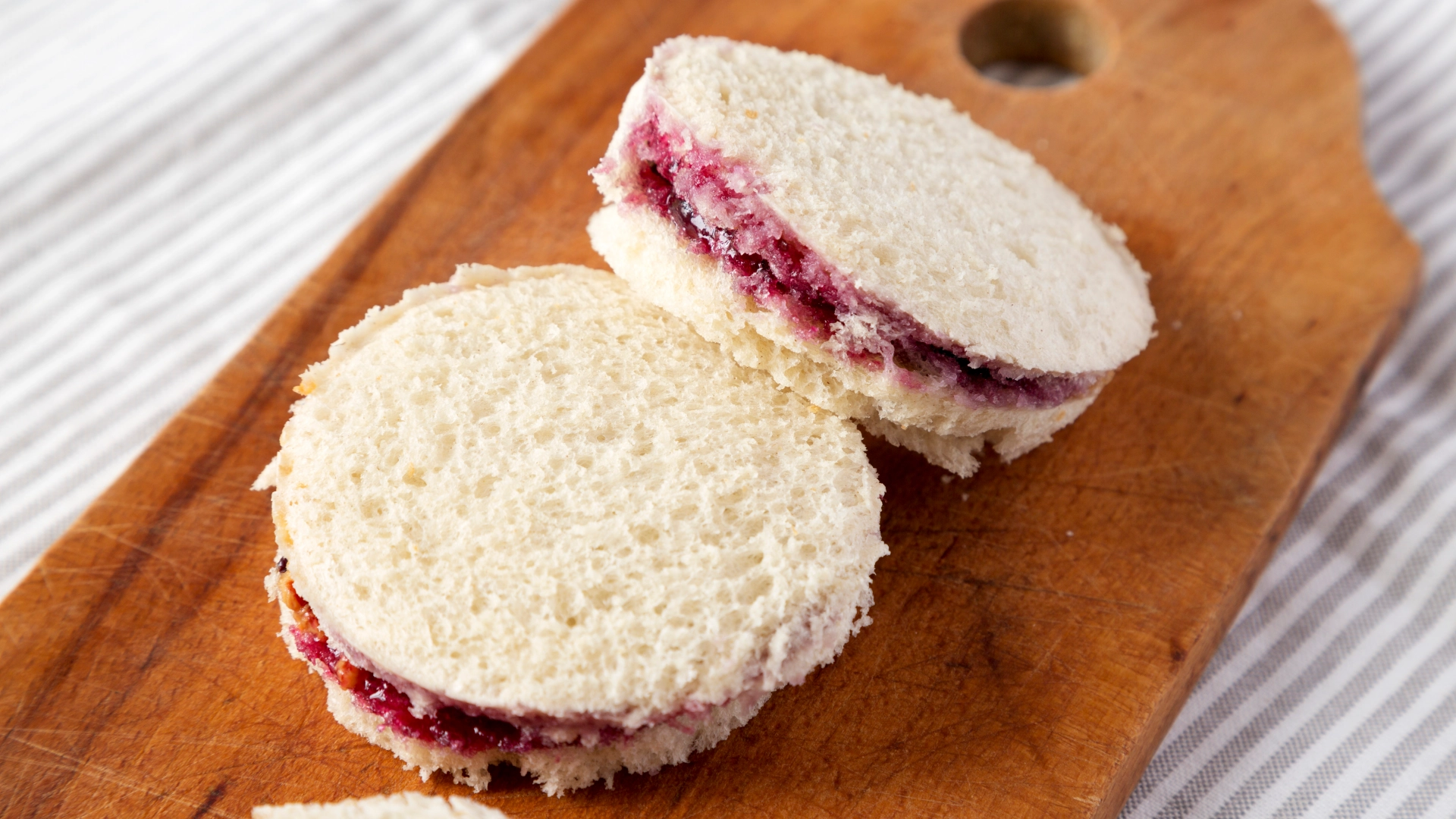 Two small, circular jam sandwiches made with Hovis Soft White Medium Sliced placed on a wooden chopping board, with the jam slightly visible between the slices of bread.