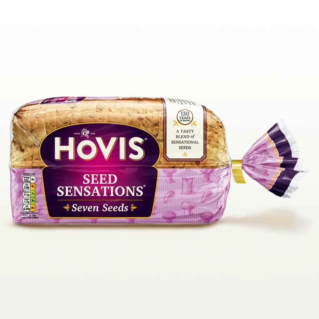 Hovis Seed Sensations Original bread on a white backdrop with a subtle shadow. The packaging is half transparent and half pink, decorated with images of flour, a chef's hat, seeds and wheat.