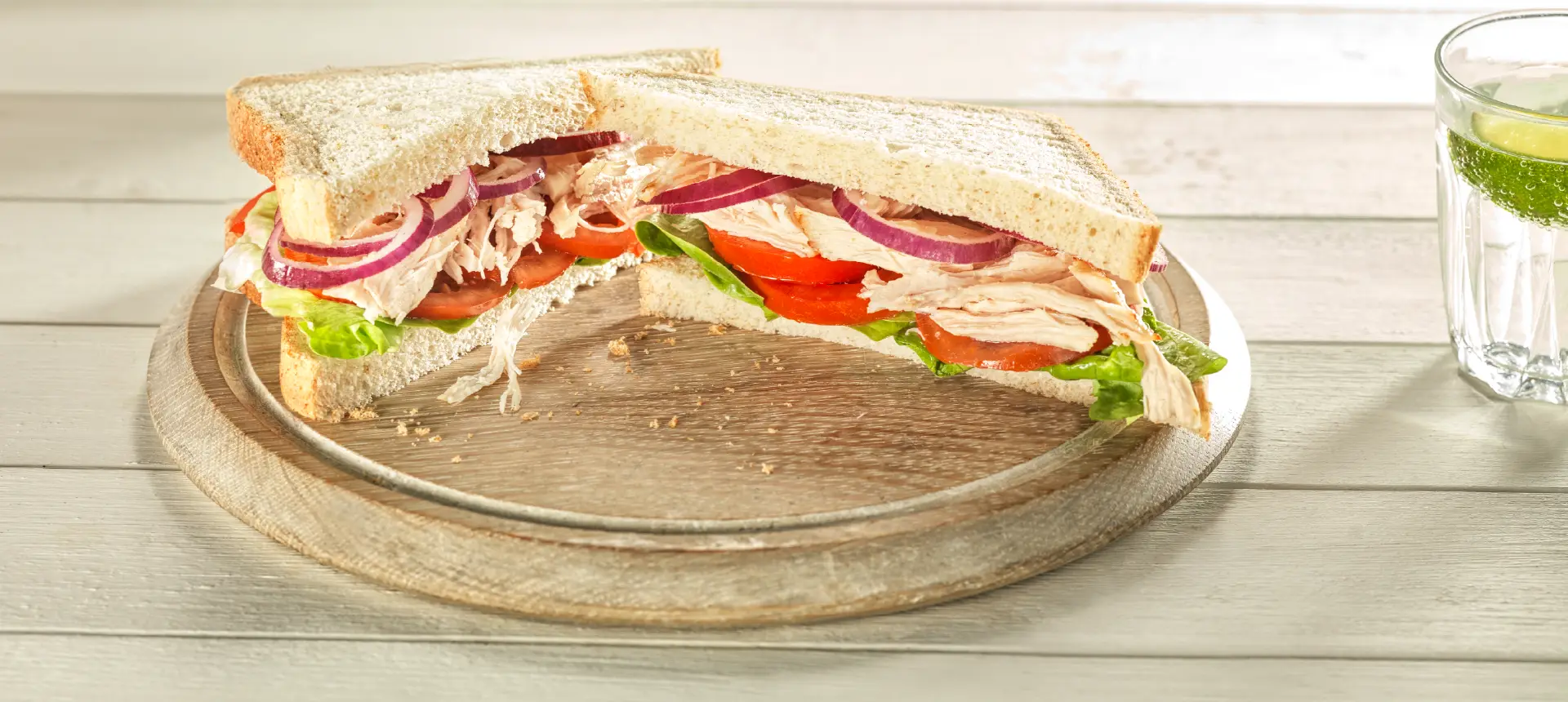 Two halves of a chicken sandwich made with Hovis Best of Both bread, filled with red onions, lettuce, and tomatoes, on a wooden board.