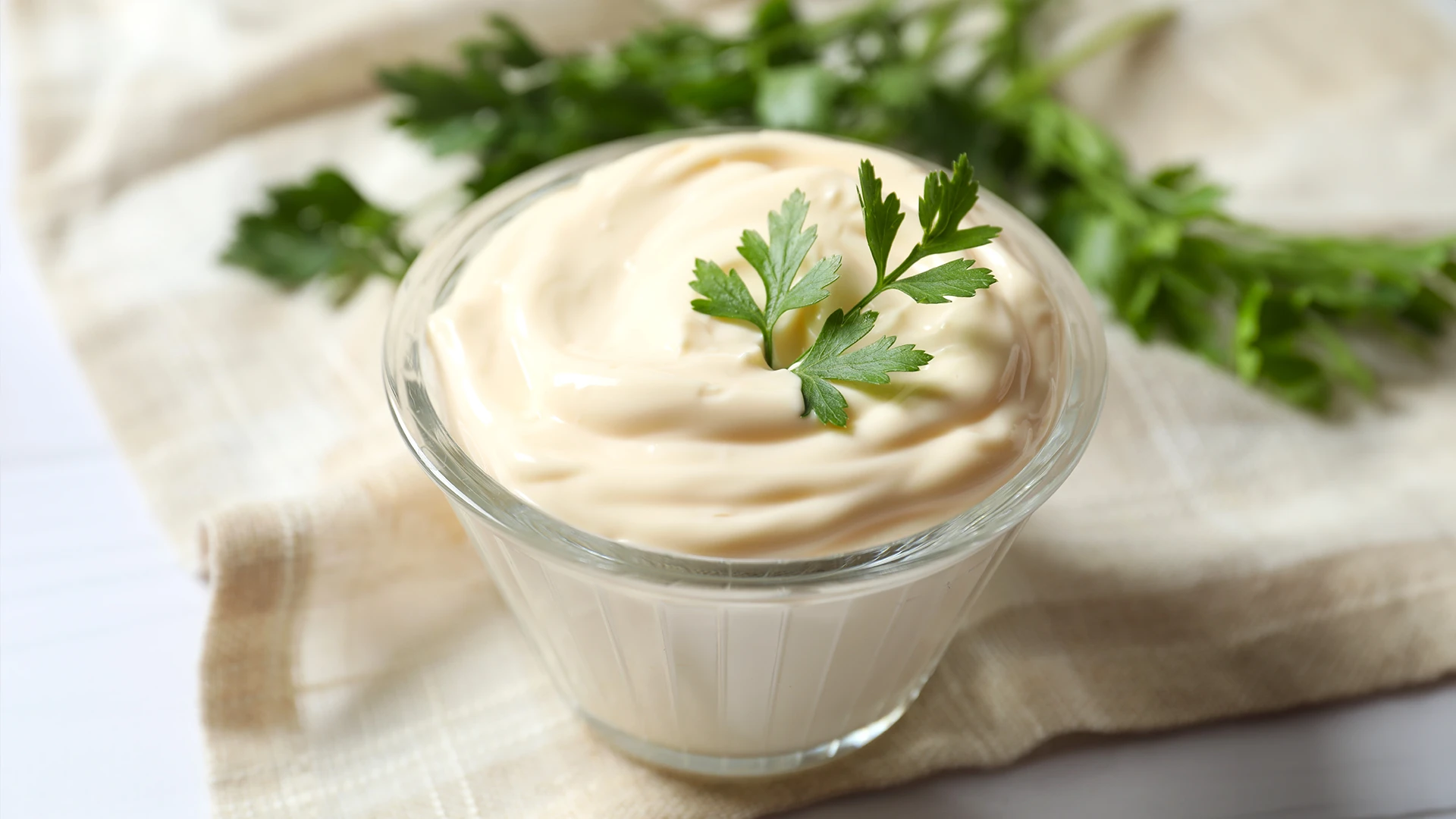 A small glass bowl filled with mayonnaise with a parsley leaf on top, resting on top of linen napkins. 
