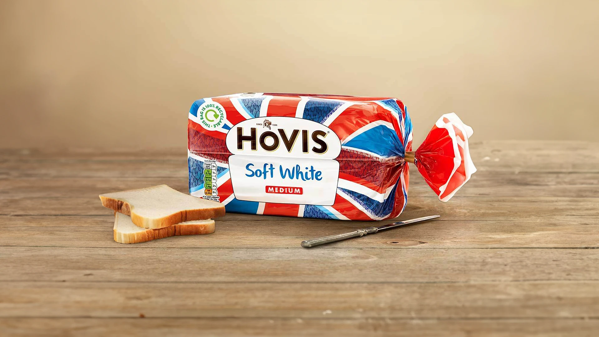 Hovis Soft White Medium Sliced bread inside its Union Jack-inspired packaging, with two slices of bread and a metal knife beside it.