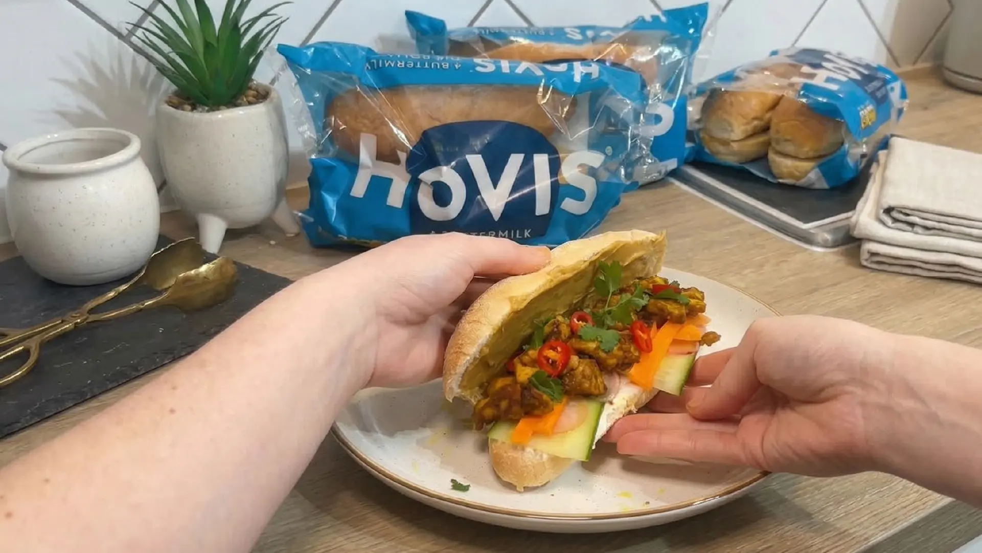 Two hands hold the open sub roll on a white plate. The sub roll is loaded with satay chicken and pickled carrots and radishes. 