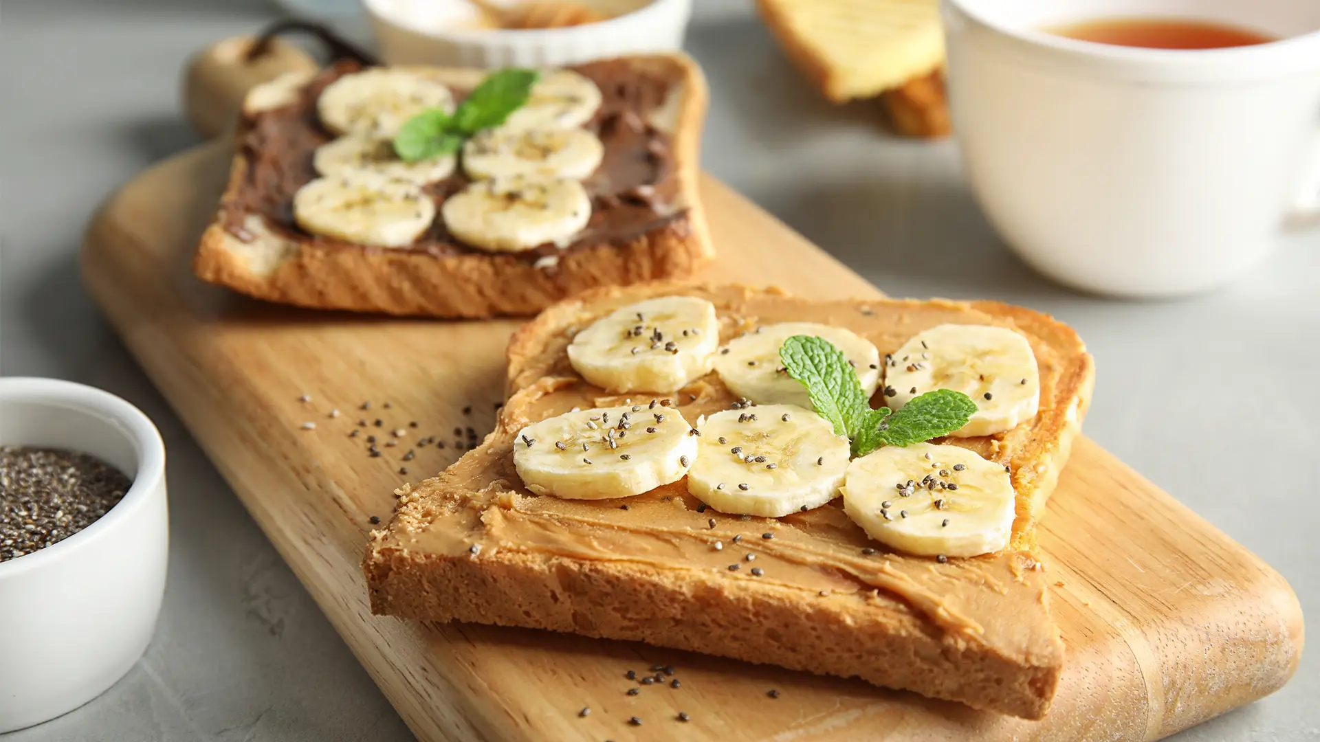 A top-down shot shows two slices of toast on a wooden cutting board. The front slice is topped with peanut butter, banana slices, and a sprinkle of chia seeds, garnished with a sprig of mint. The back slice has a chocolate spread, banana slices, and a sprig of mint. To the left, a small white bowl holds more chia seeds. In the blurred background, a white teacup with a spoon and a slice of plain toast are visible. The surface beneath the board is a light gray.