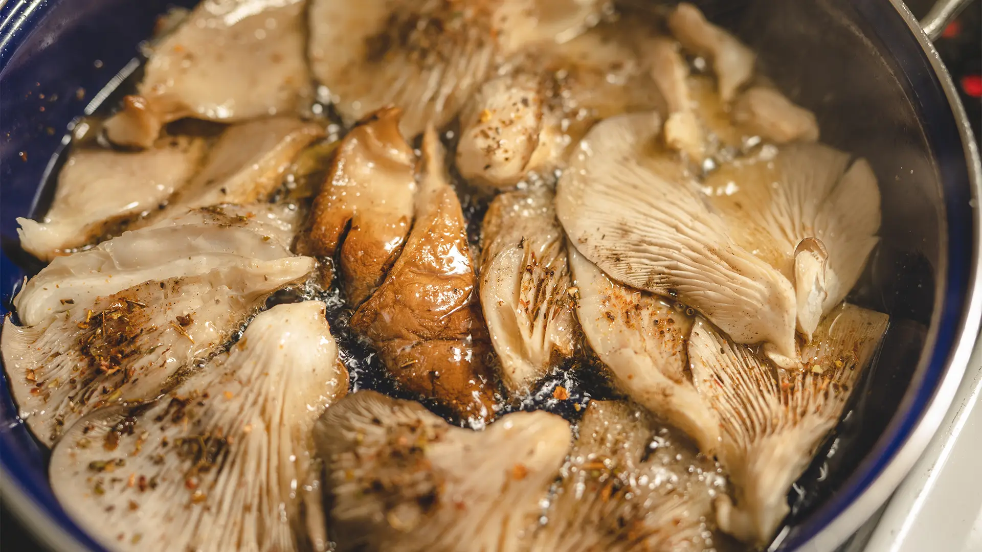 A top-down view of oyster mushrooms releasing steam as they cook in a medium-sized metal frying pan on a hot stovetop.