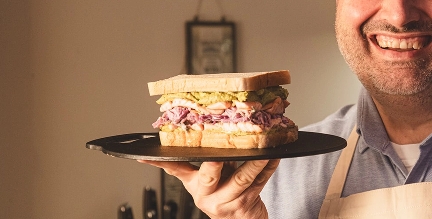 A sandwich made with Hovis Soft White, smashed avocado, seared salmon and coleslaw is being held up by Wynne Evans as he smiles. 