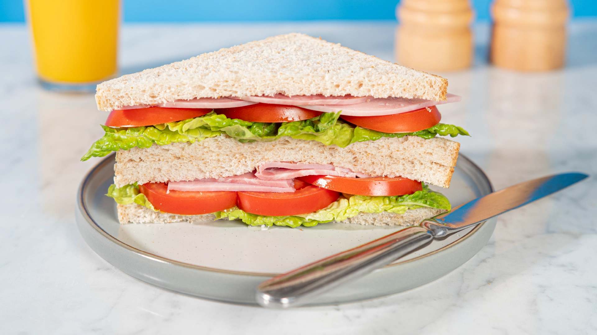 Two halves of a Hovis Best of Both Medium Sliced sandwich, stacked and filled with thin slices of ham, tomatoes, and lettuce leaves, on a plate with a butter knife on the side.