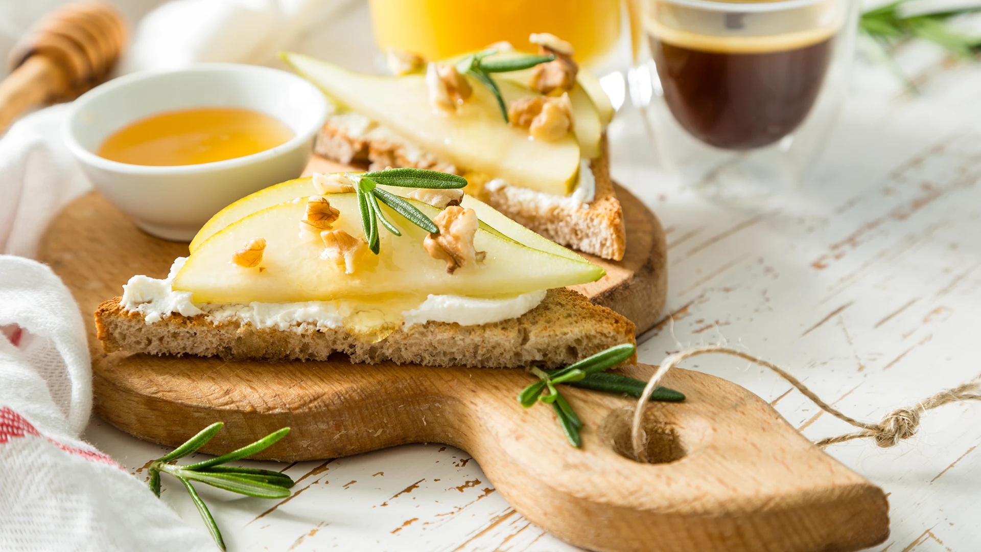 A halved slice of Hovis Nimble Wholemeal topped with ricotta, two thin pear slices, and walnut pieces on a wooden board, with a saucer of honey in the background.
