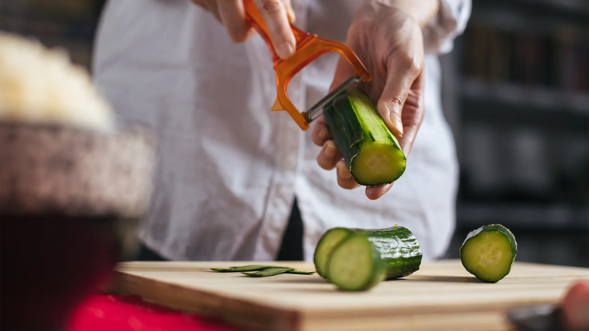 A person holding an orange vegetable peeler in one hand, and half a cucumber on the other, cutting thin slices of cucumber on a wooden cutting board.
