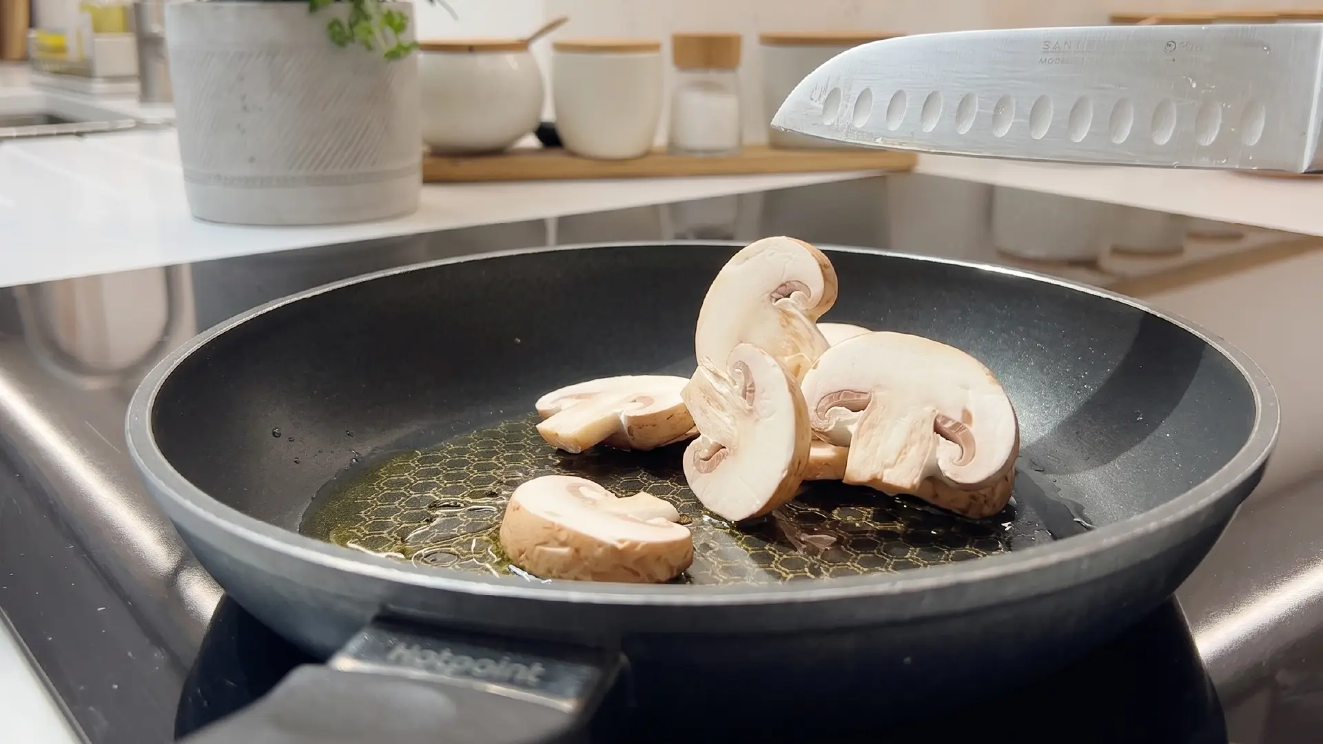 Sliced mushrooms being added from a knife into a black frying pan with sizzling oil on a stovetop.