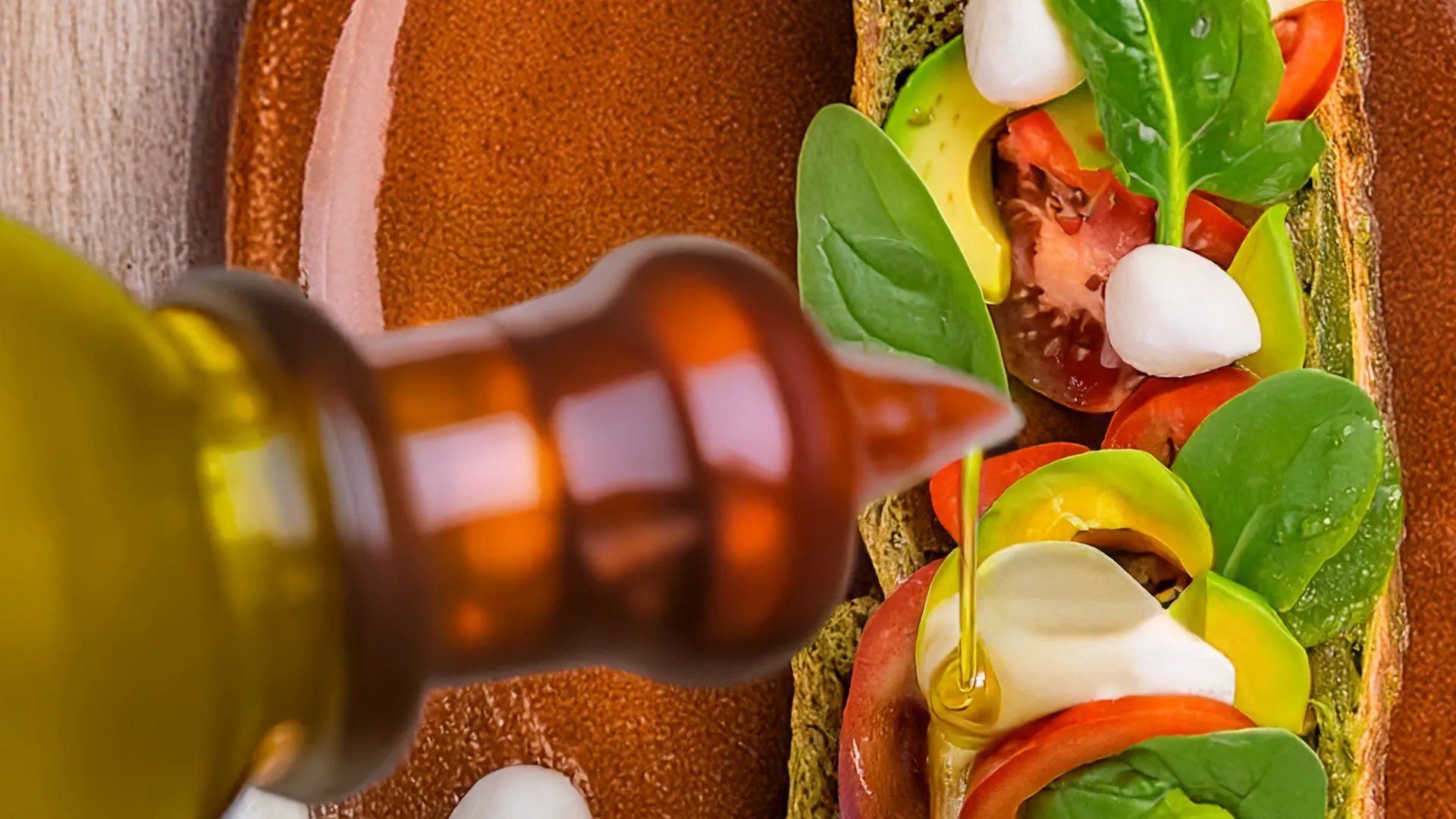 the bottom half of a sub roll is on the plate with tomato, spinach, avocado and mozzarella on top. The spout of an olive oil bottle is drizzling olive oil on top of the half sandwich. 