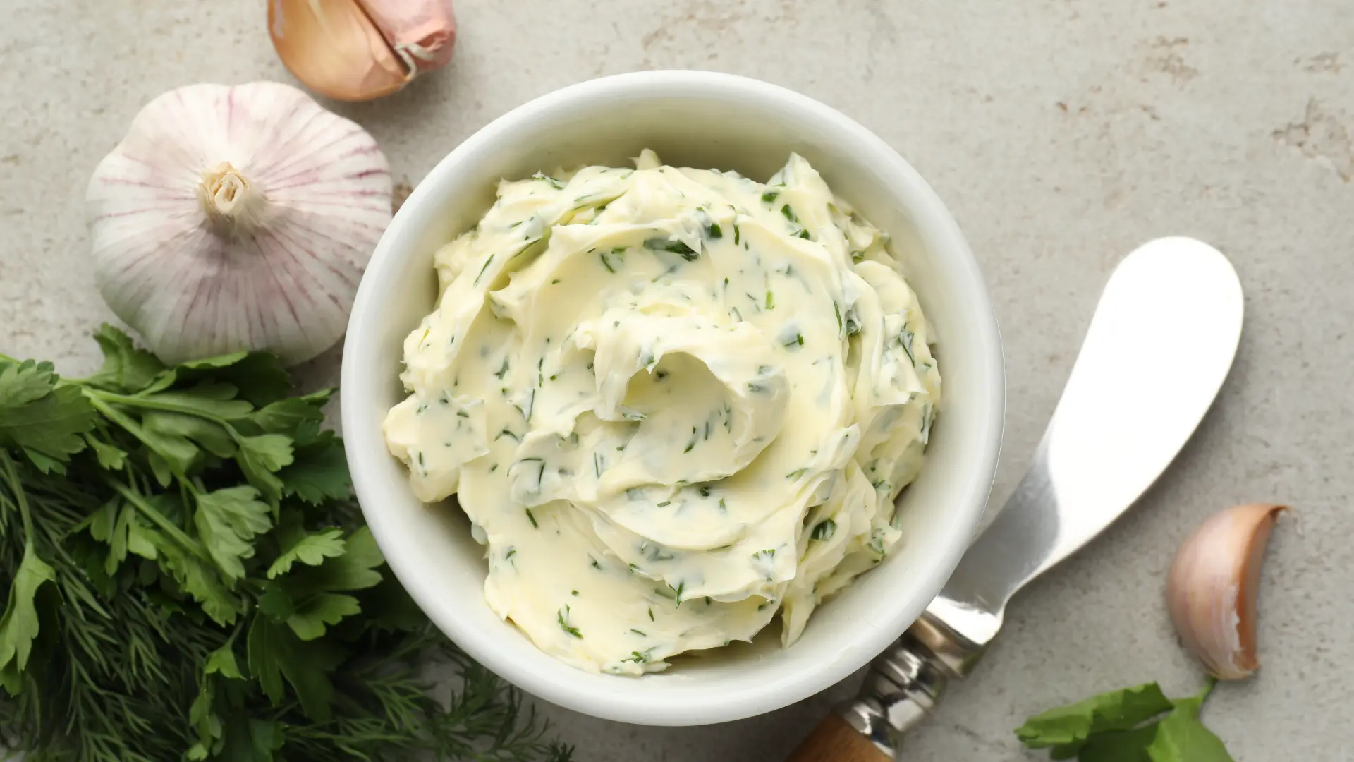 A white bowl filled with swirled herb butter, flanked by fresh parsley, garlic cloves and a silver butter knife.