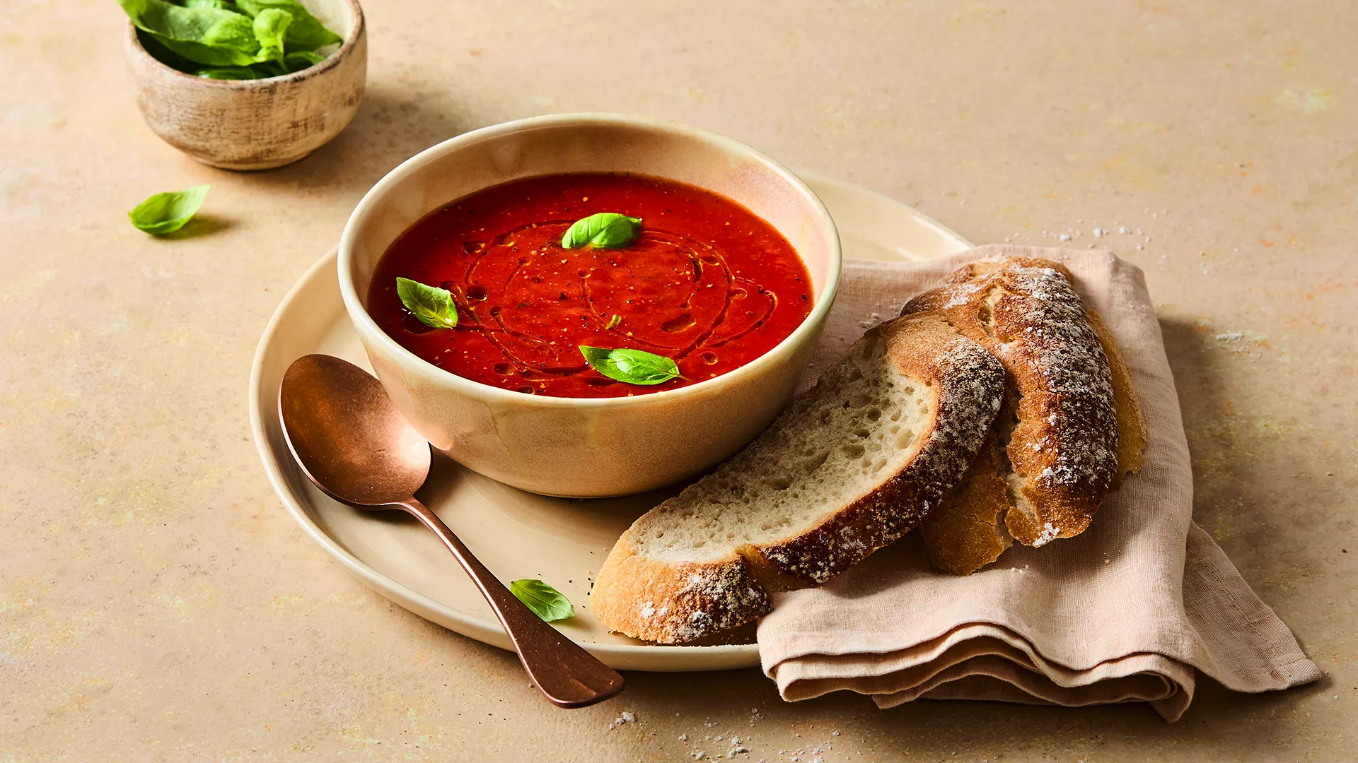 A close-up image of a Hovis®  White Sourdough slice on a plate next to a bowl of Tomato and Basil Soup sprinkled with basil leaves.