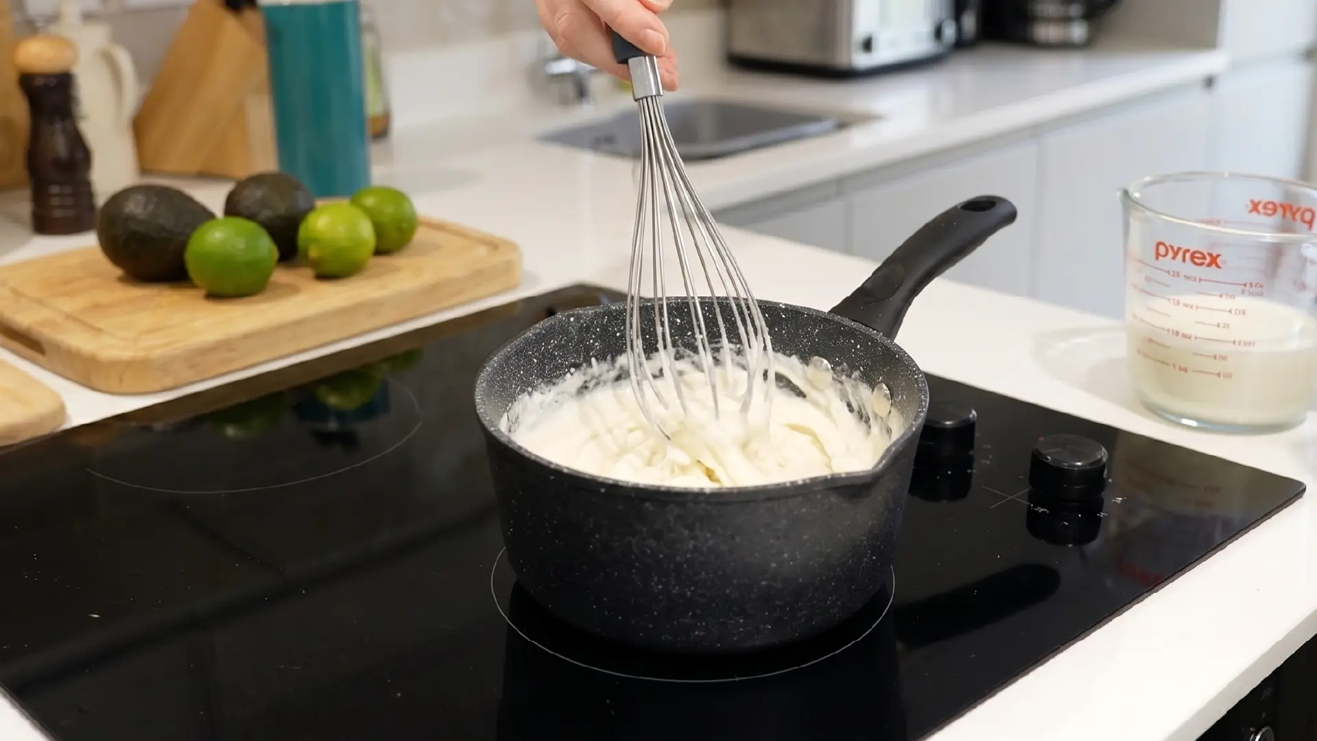 A hand whisks a creamy white mixture in a black pot on a stovetop, with avocados, limes and a measuring cup nearby.